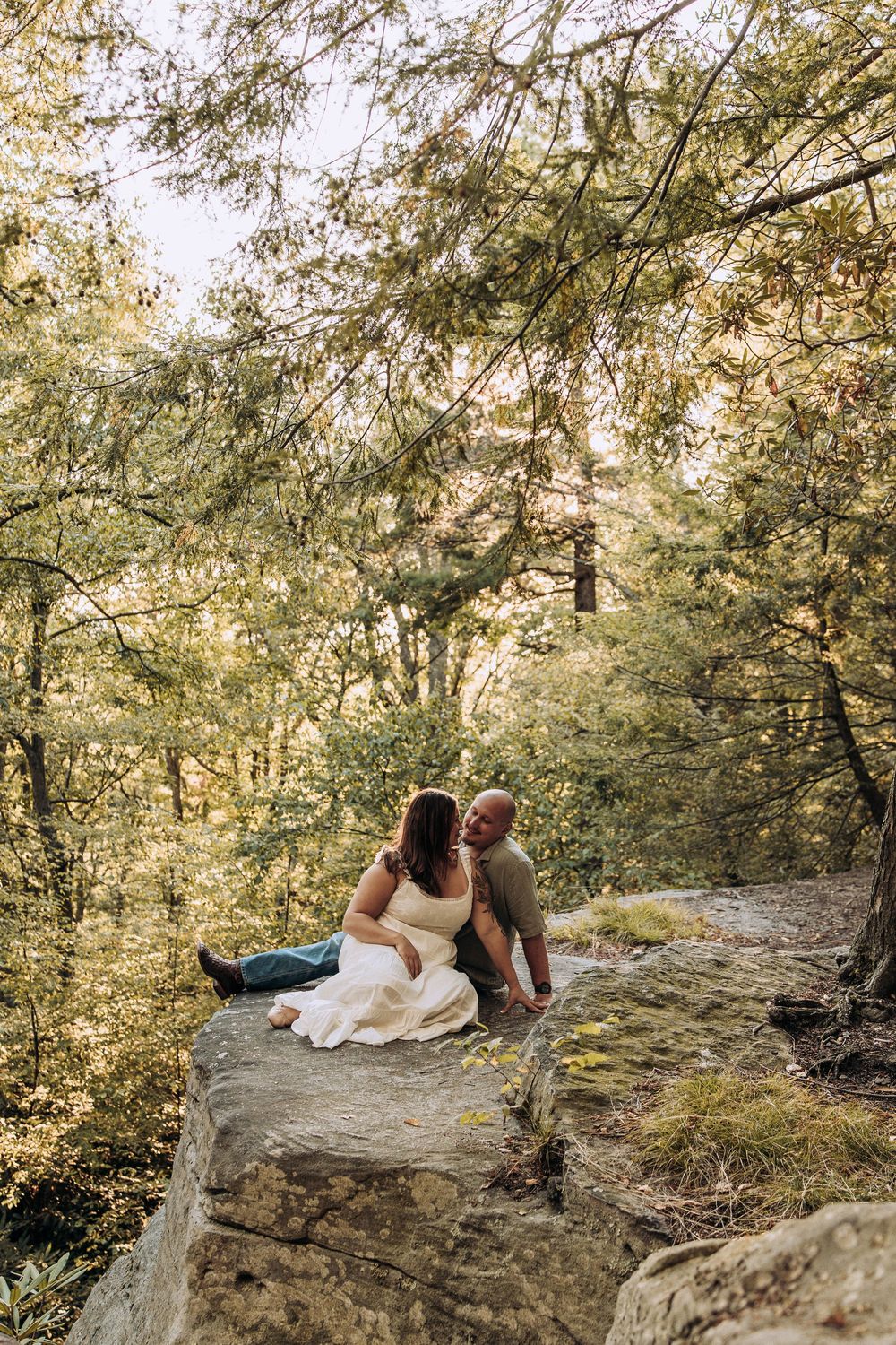 Couple sitting closely together on a large rock surrounded by lush forest trees.