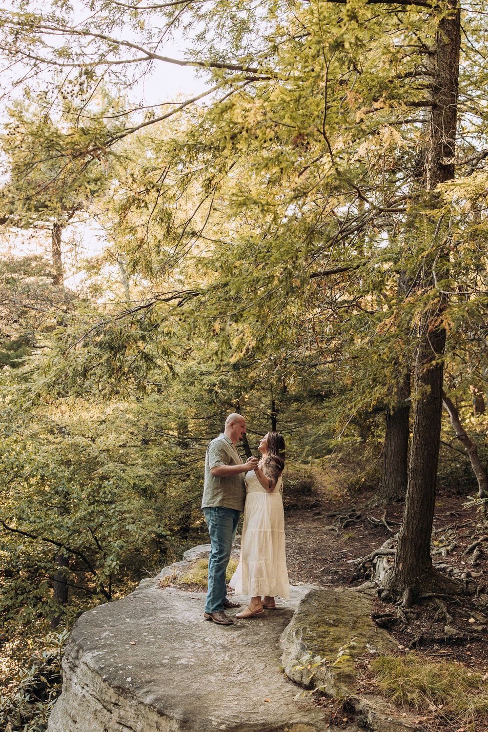 Couple standing together on a stone path surrounded by tall green trees in a forest.