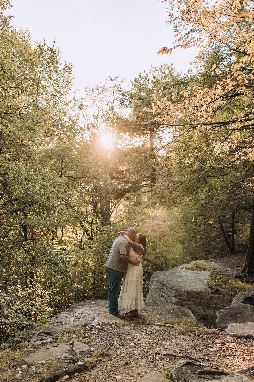 Couple sharing a romantic kiss on rocks with golden sunlight filtering through trees.