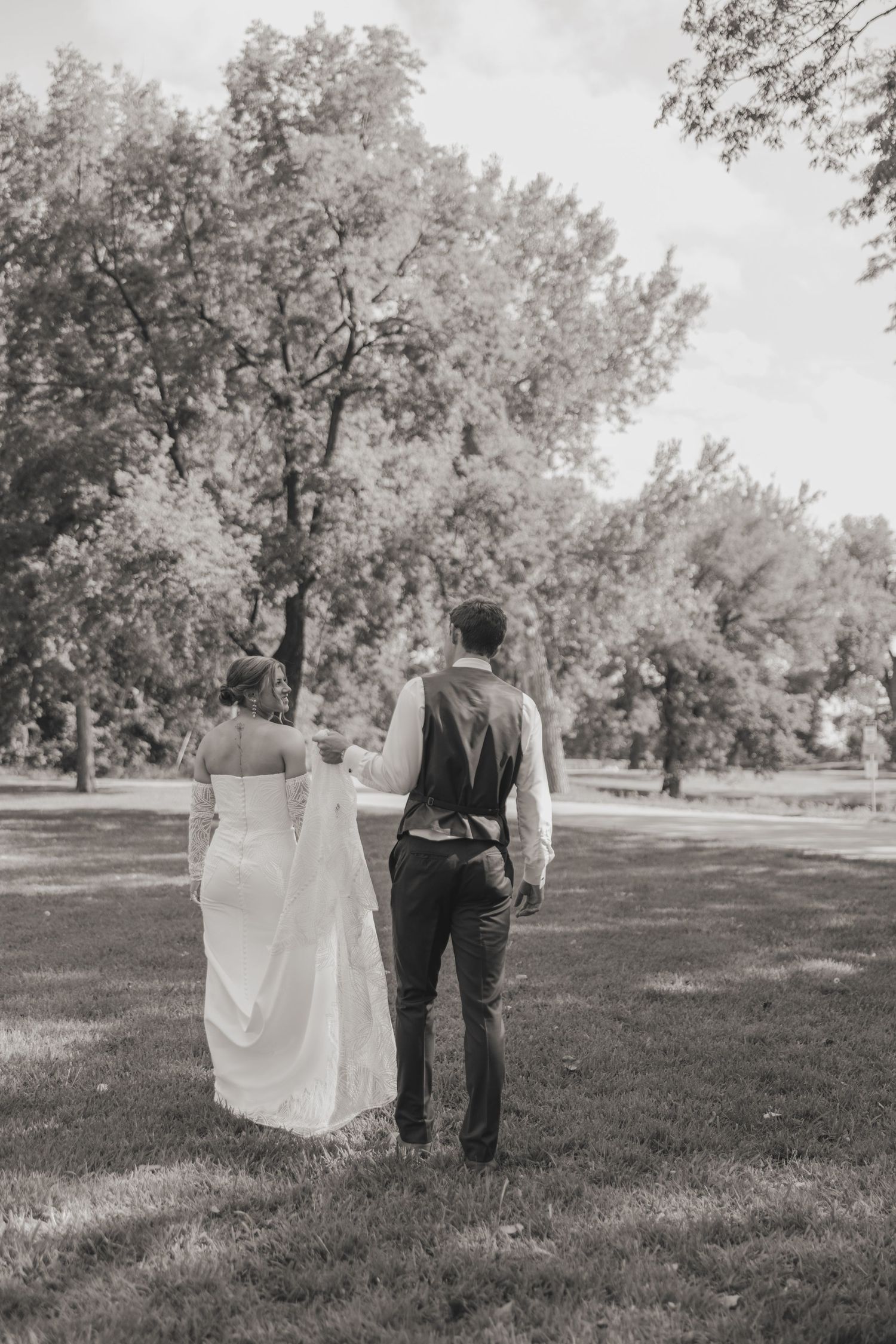 A couple in wedding attire walks away from the camera through a serene park setting in black and white photography.