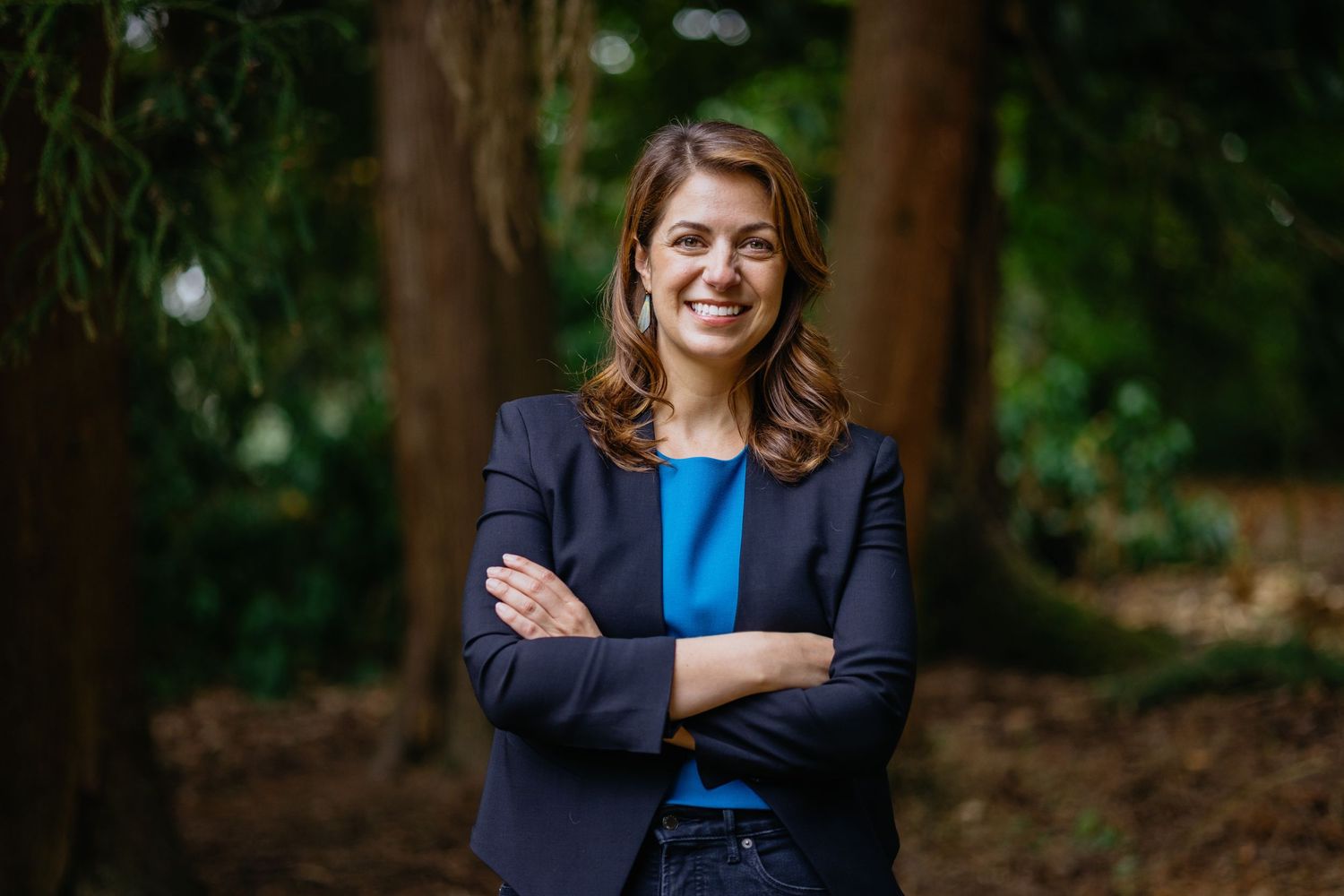 Professional woman in navy blazer and blue top smiling with arms crossed outdoors among green trees.