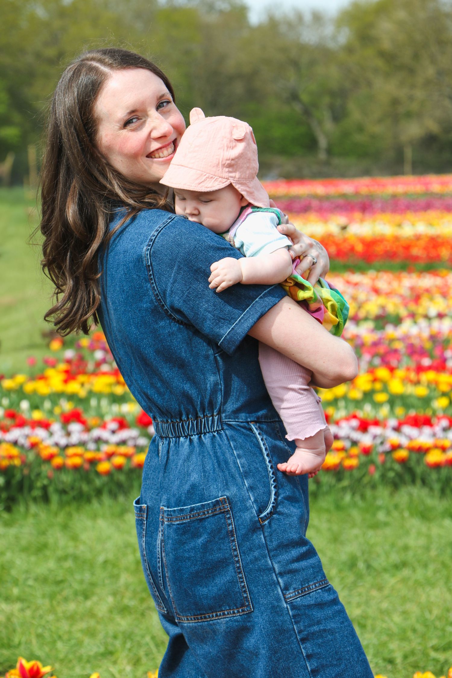 A person in denim dress holds a baby while standing in a vibrant tulip field on a spring day.
