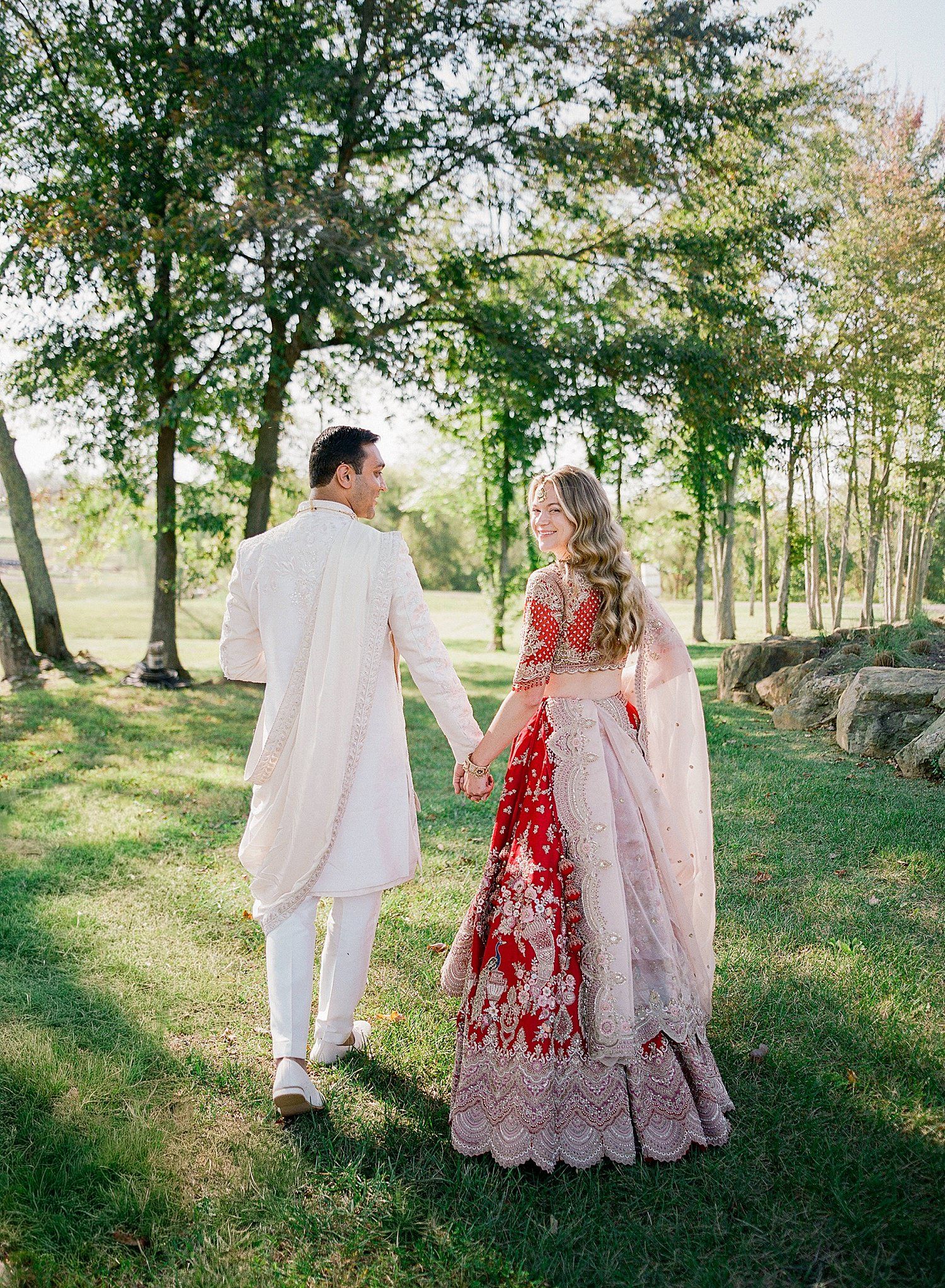 A couple in white and red traditional attire walking through a scenic wooded area during a summer photoshoot.