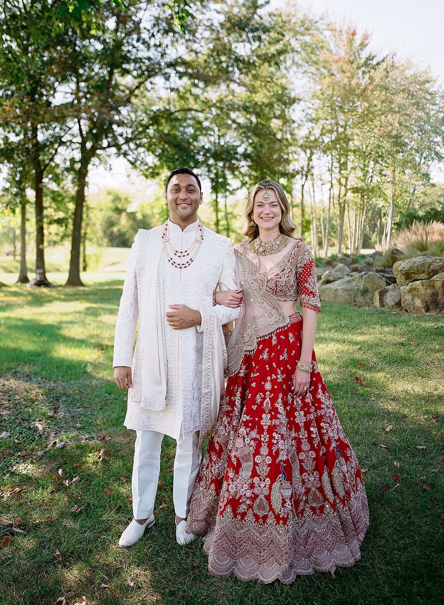 A couple poses in traditional wedding attire featuring a red and silver lehenga and white sherwani in an outdoor garden setting.