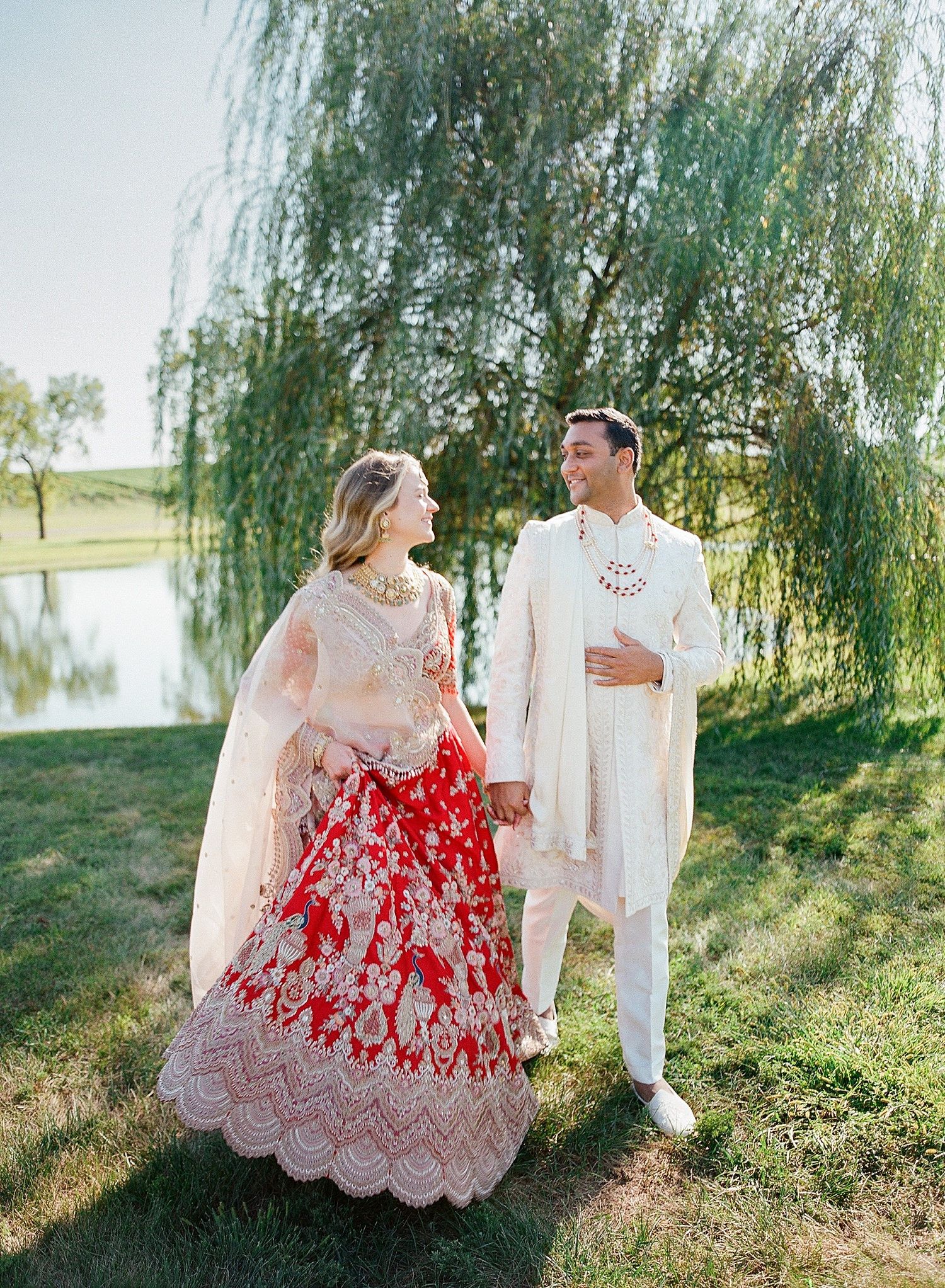 A couple in white and red lace attire poses romantically under a weeping willow tree during an outdoor photo session.