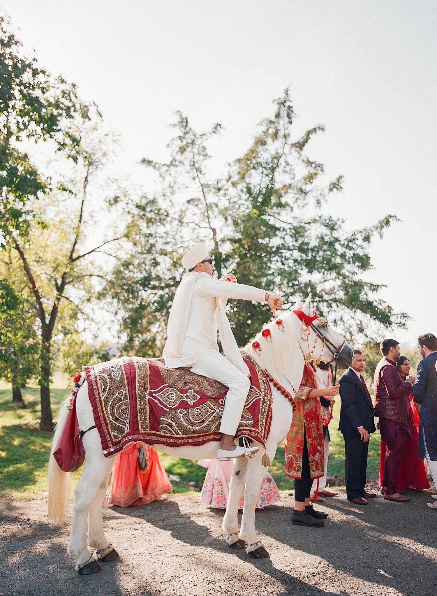 Wedding party members in burgundy and white traditional attire walk together on a sunlit path during outdoor celebration.