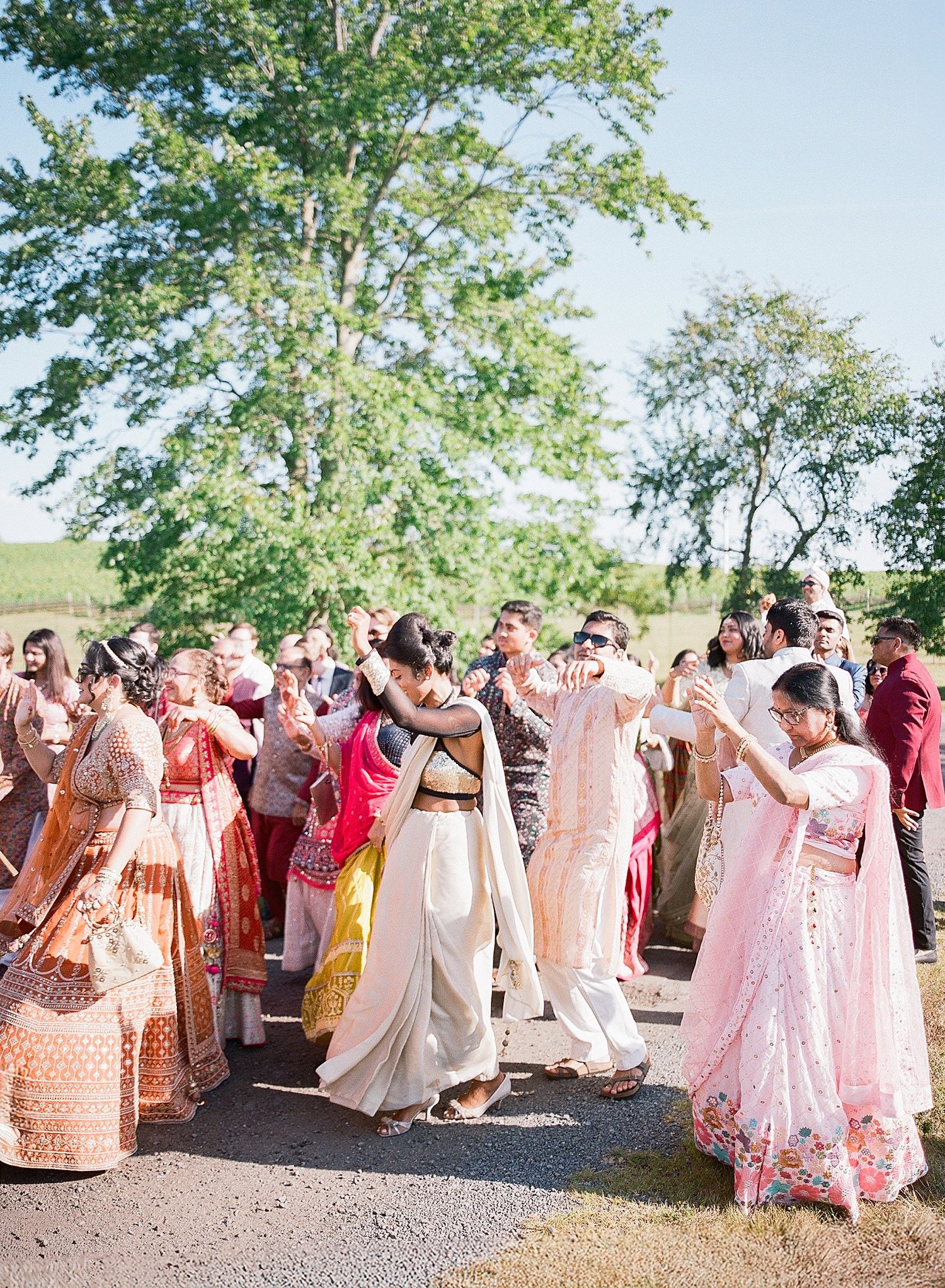 A large wedding party wearing pink and coral colored outfits gathers outdoors for a group photo on a sunny day.