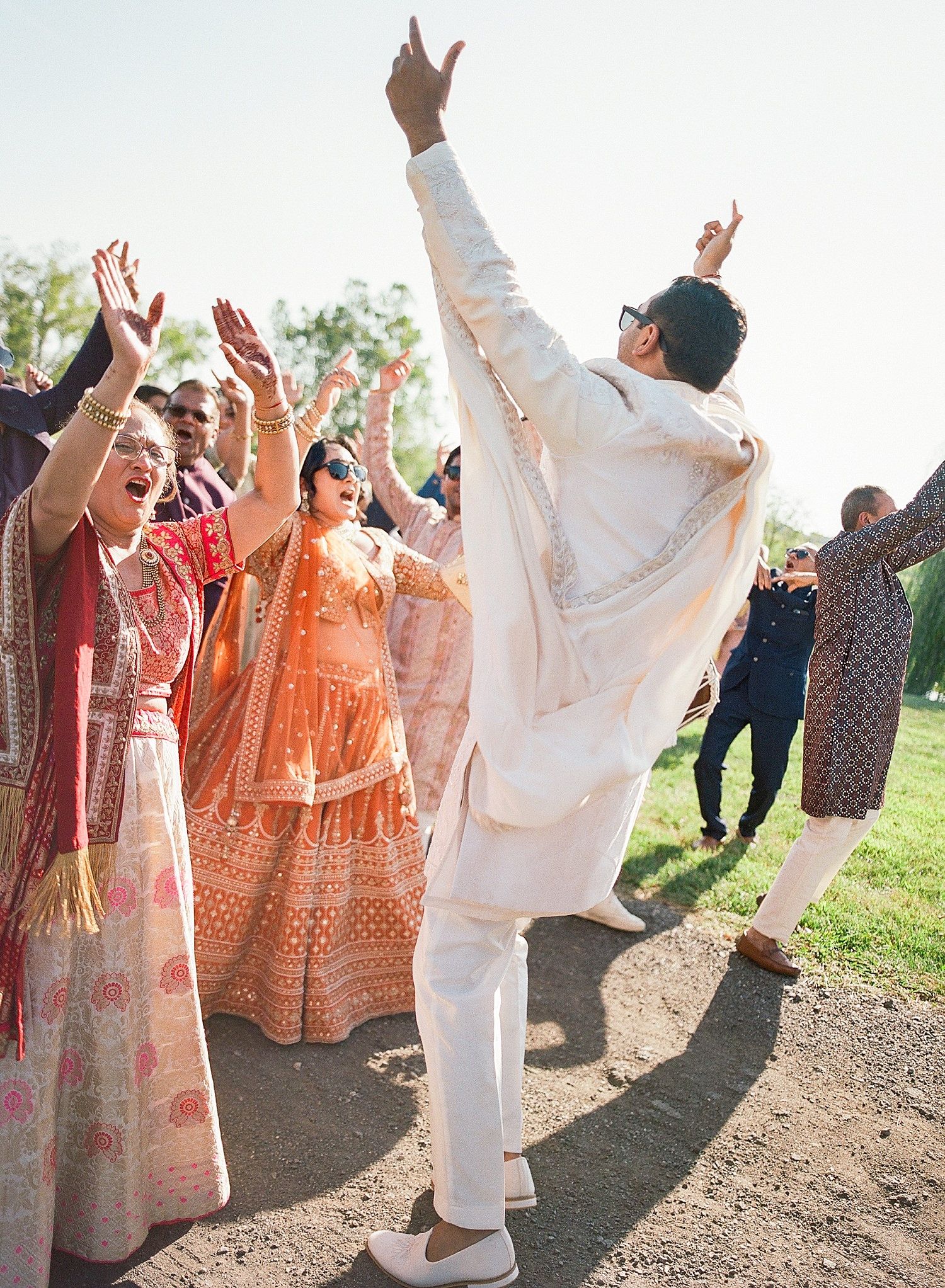 Wedding guests in traditional Indian attire dance joyfully outdoors during a celebration.