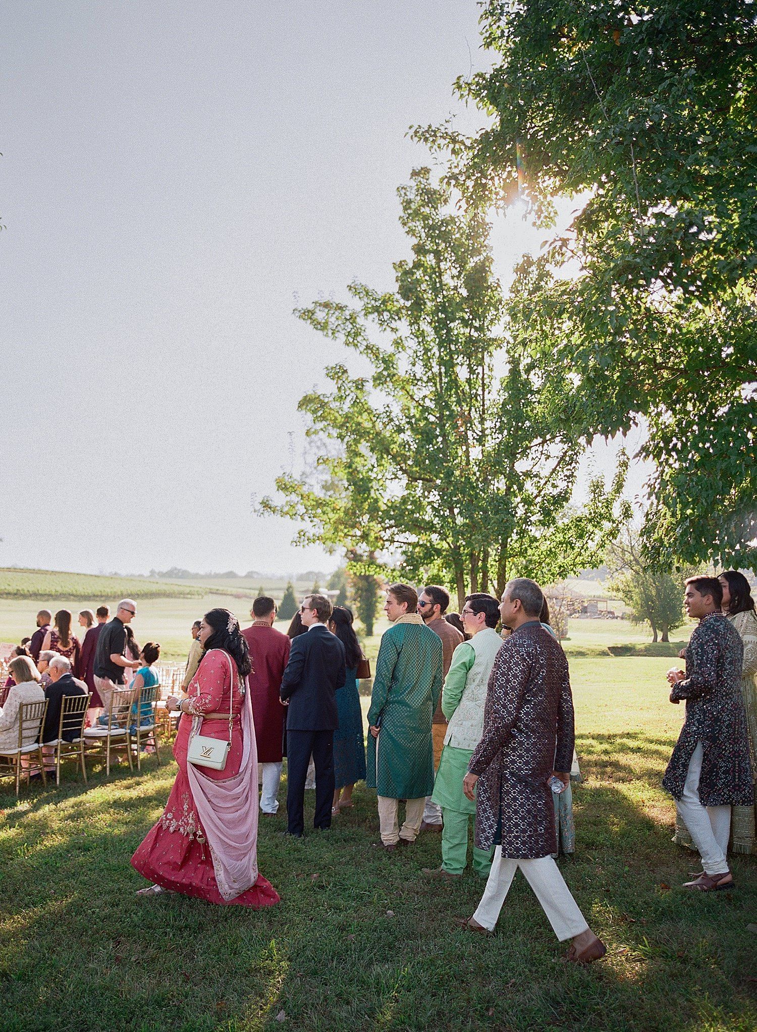 A group of wedding guests gather on a grassy hillside during an outdoor ceremony at sunset.
