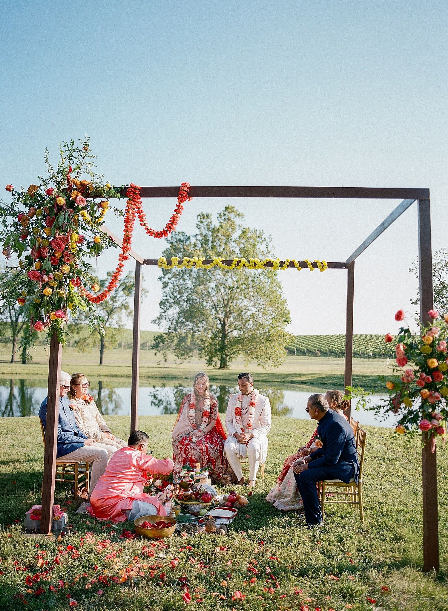 Outdoor gathering with wooden pergola decorated in red and yellow floral arrangements by a scenic lakeside setting.