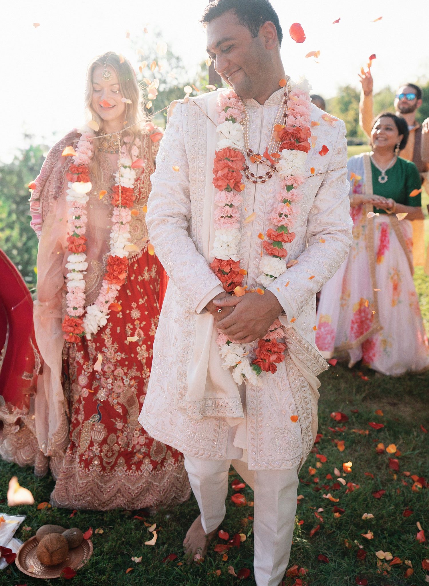 Couple in white outfits celebrates with vibrant coral flower petals being tossed around them in an outdoor garden setting.