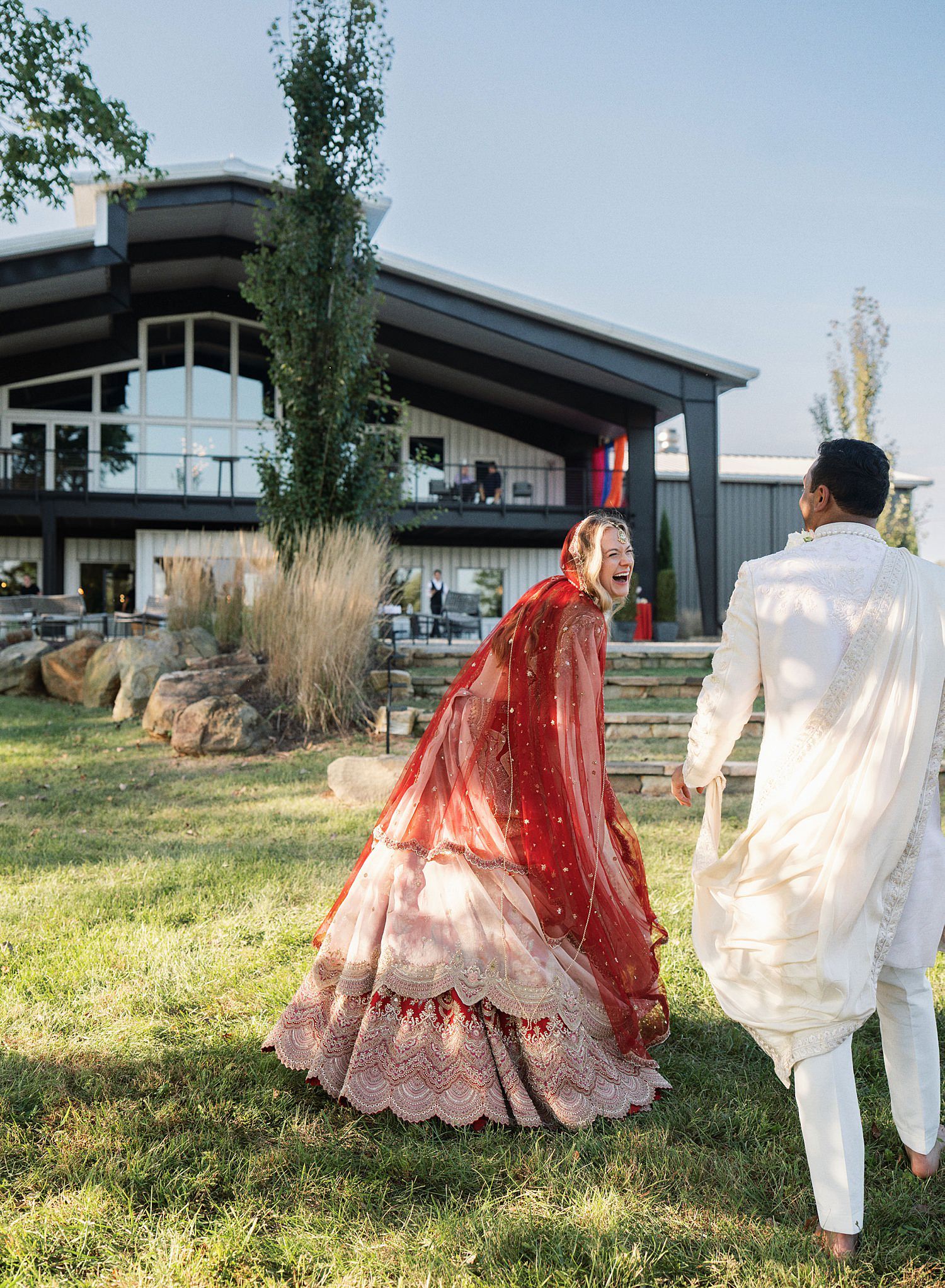 A couple in traditional wedding attire walks together on grassy grounds with modern buildings in the background.