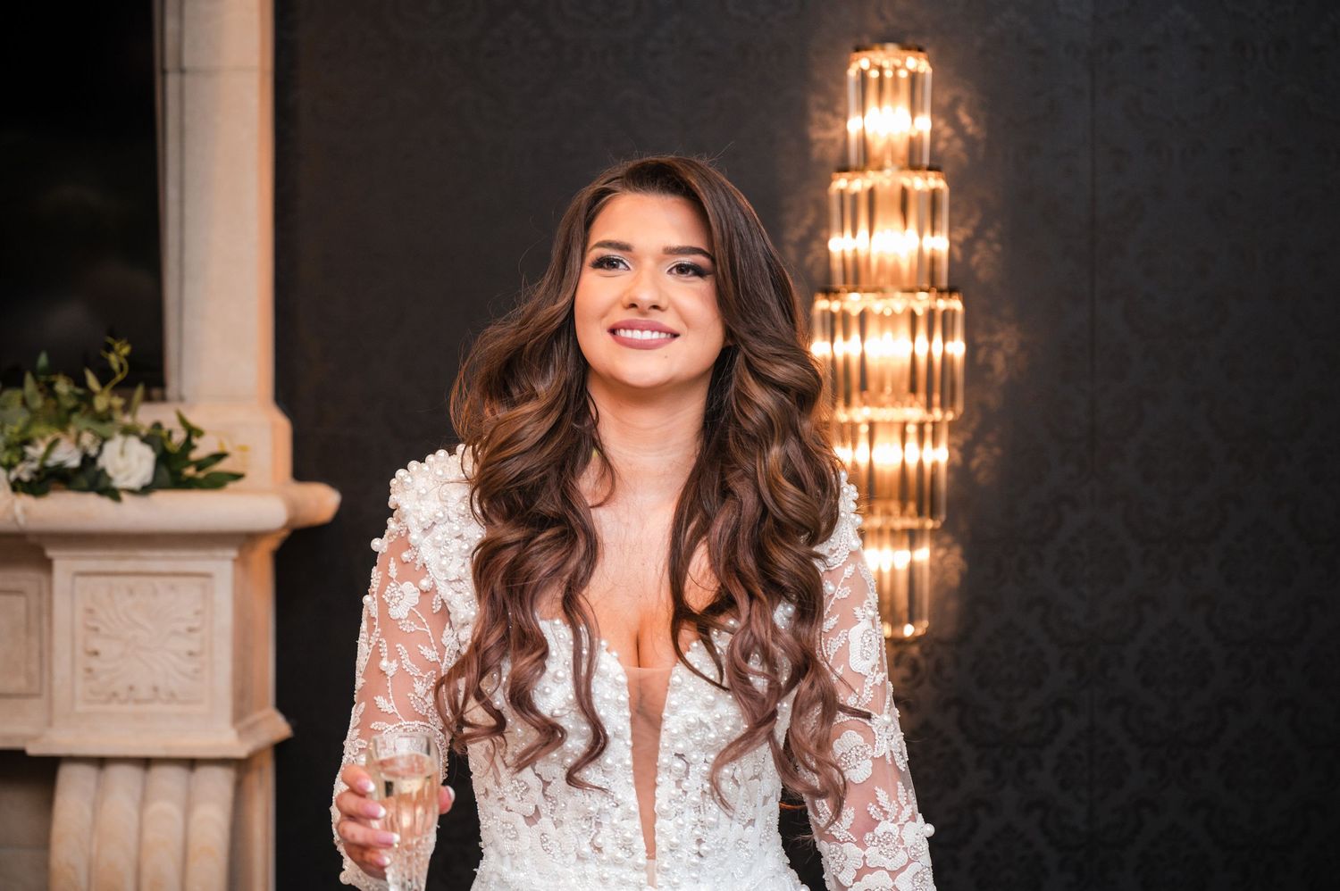 Elegant woman in white lace dress poses in front of ornate chandelier and stone columns in luxurious setting.