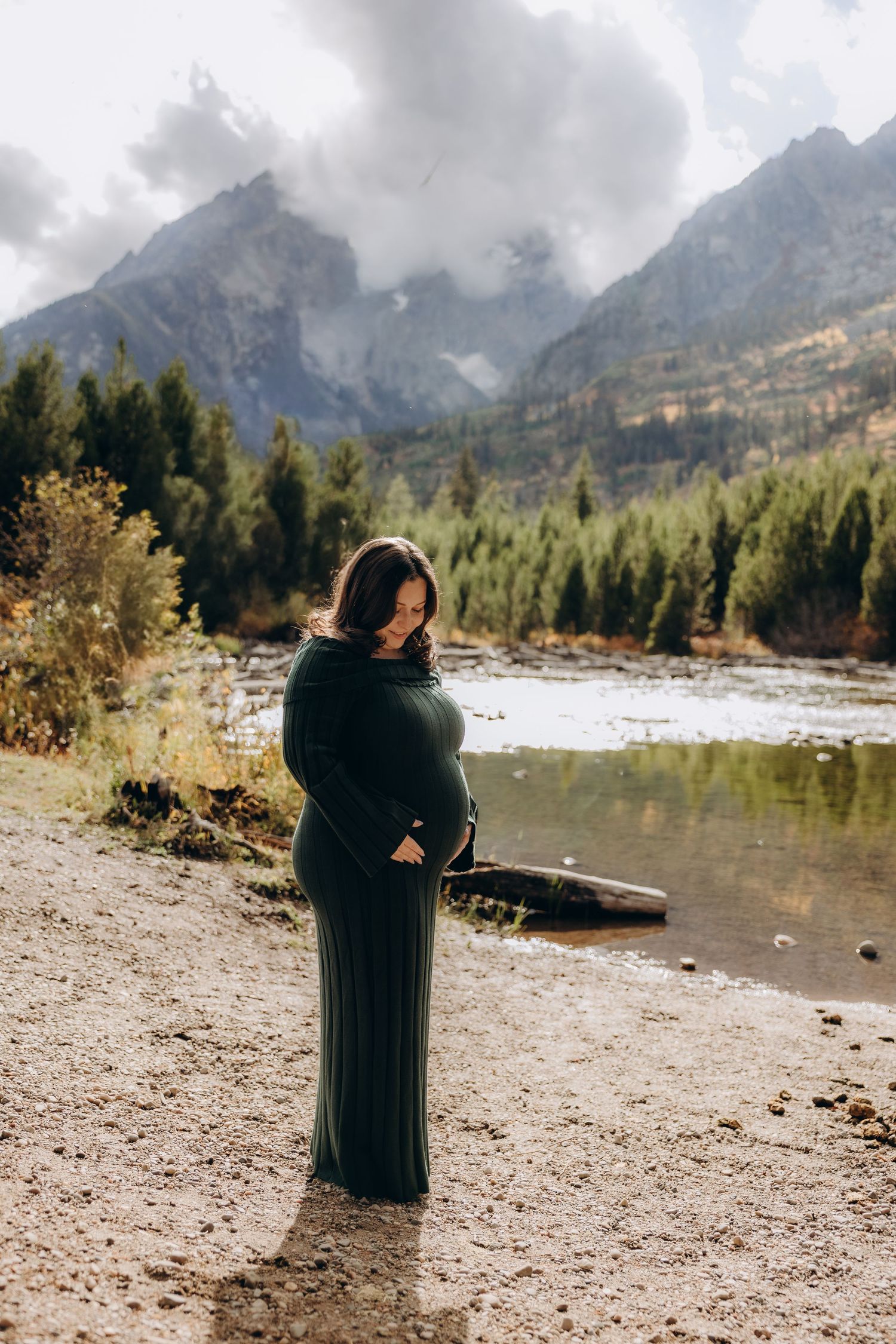 A figure in dark clothing stands on a rocky shoreline with snow-capped mountains and evergreen forest in the background.