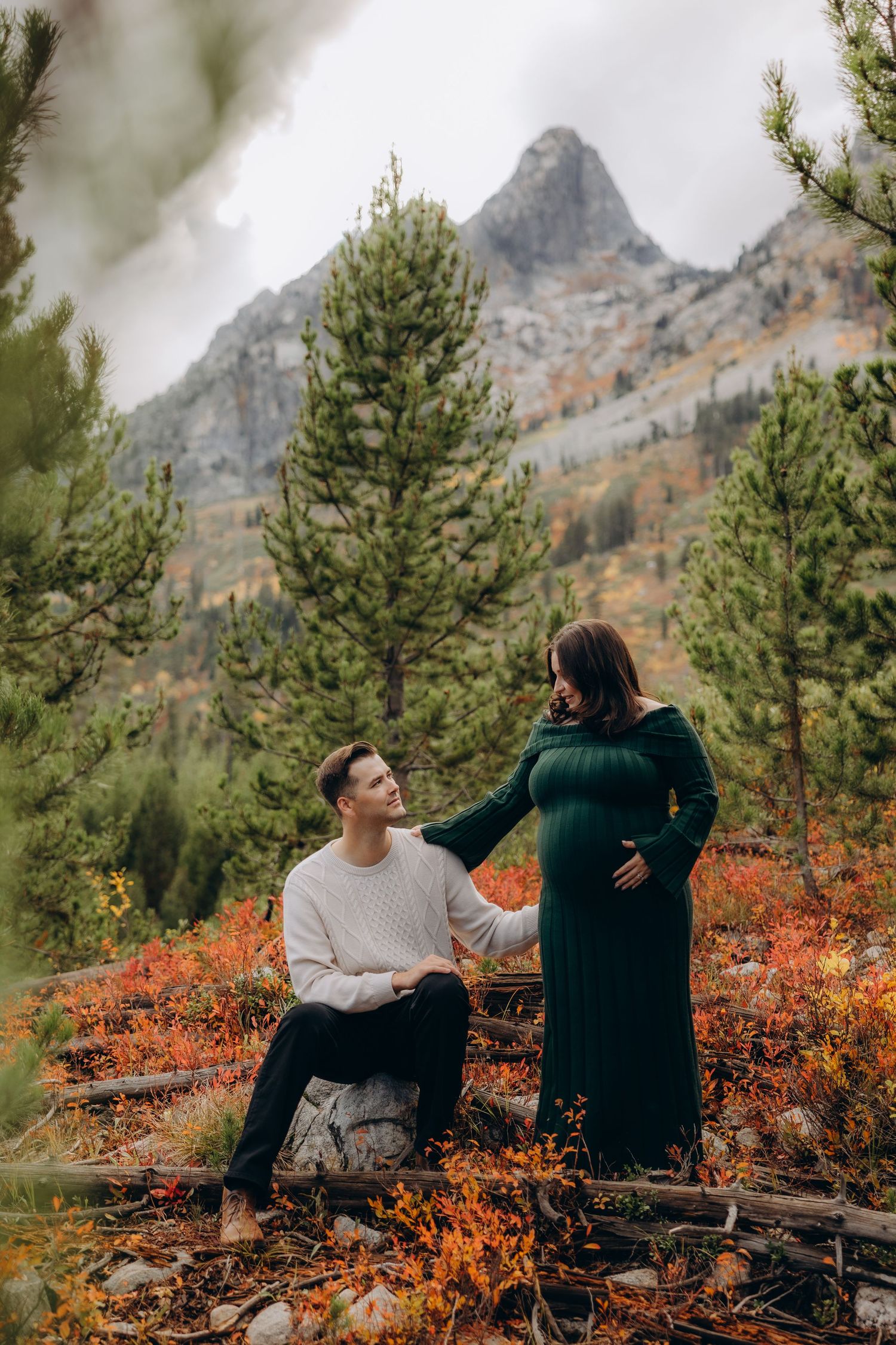Two people sit together on a mountain ledge surrounded by evergreen trees and vibrant orange foliage in autumn.