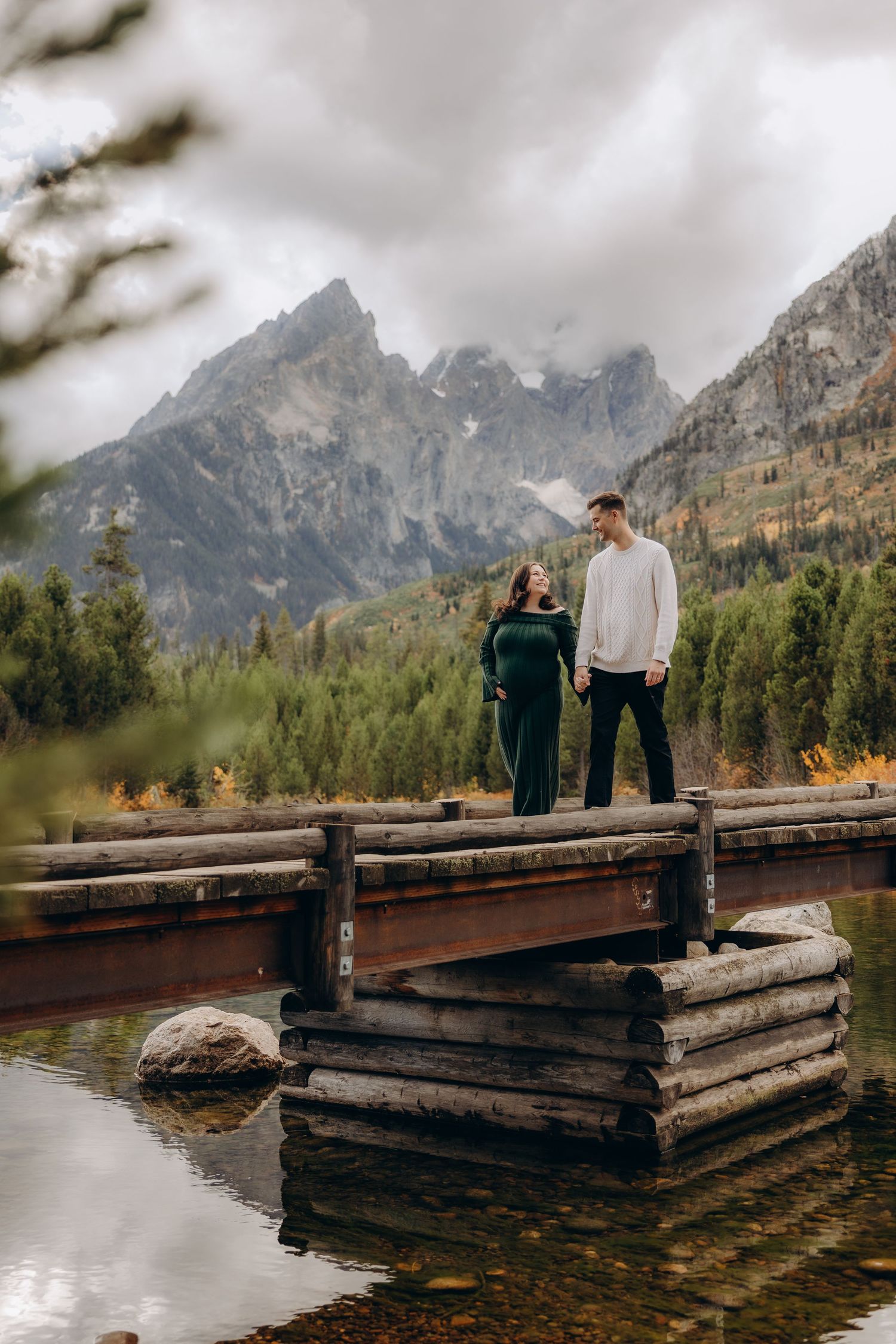A couple stands on a rustic wooden dock overlooking a lake with dramatic mountain peaks in Grand Teton National Park.