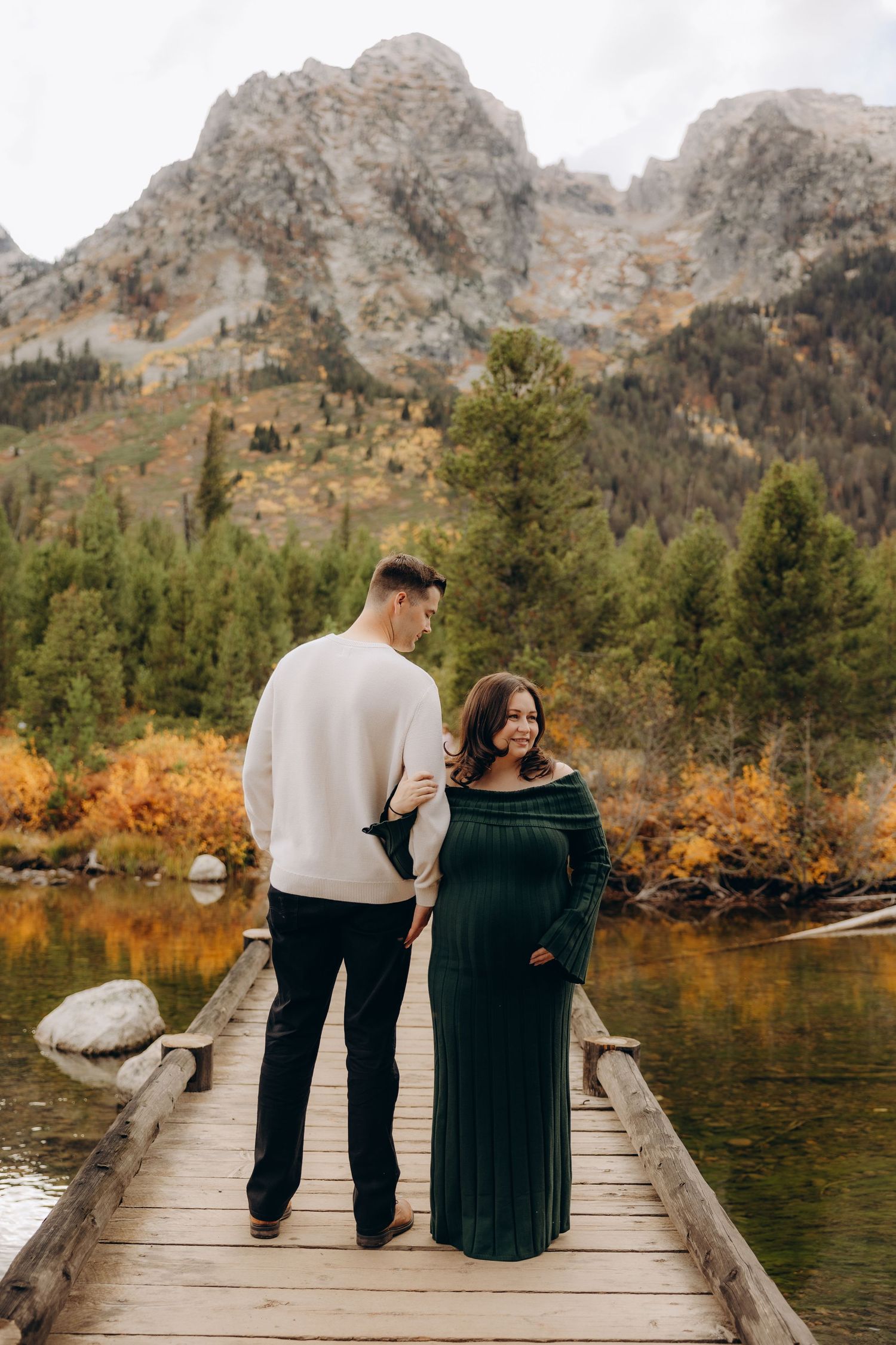 A couple stands together on a wooden dock overlooking a serene mountain lake surrounded by autumn foliage.