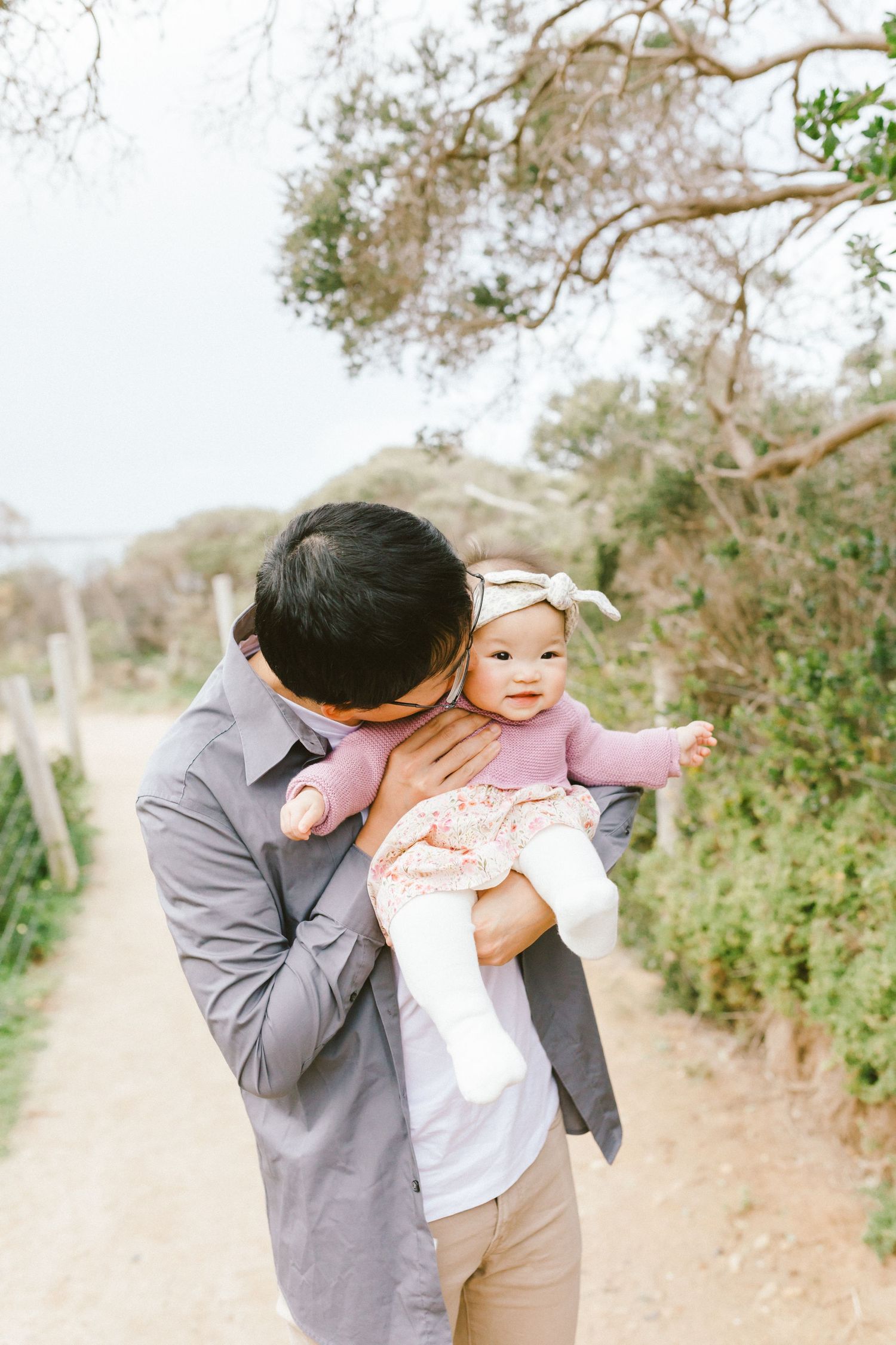 A parent in a gray sweater lifts and plays with a small baby wearing pink outdoors on a sunny day.