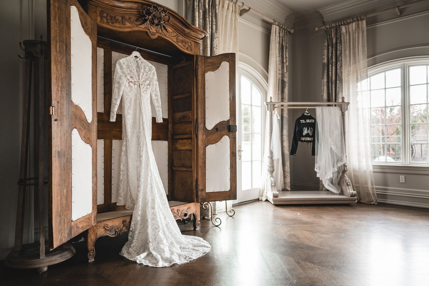 An elegant lace wedding dress hangs from an ornate wooden wardrobe in a sunlit dressing room.