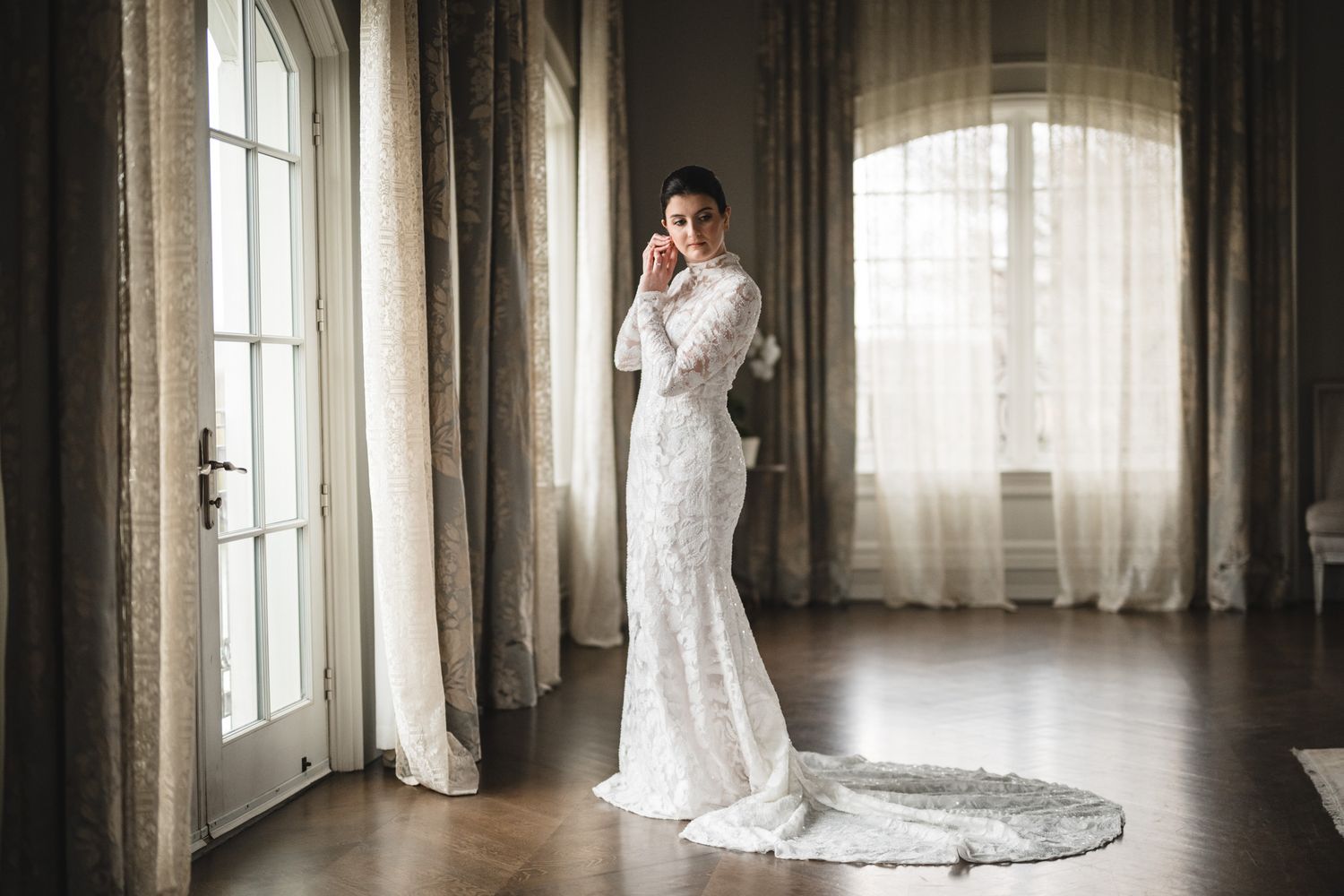 Elegant lace wedding gown with long train photographed in a light-filled room with flowing curtains.