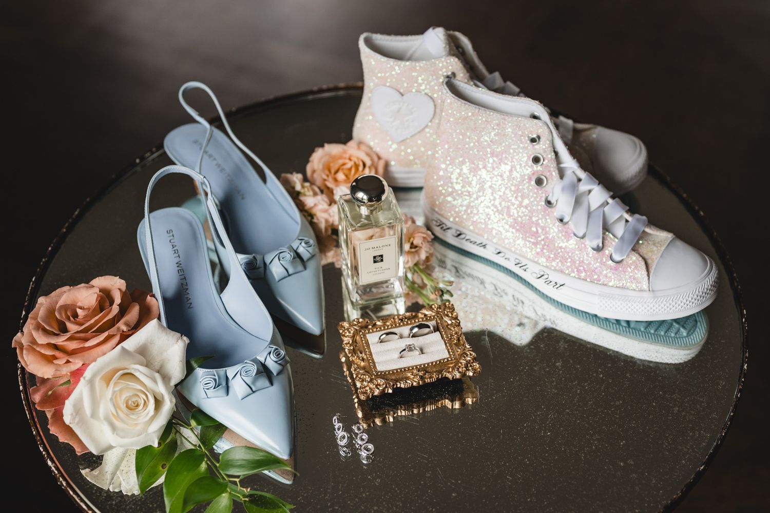 Wedding accessories including blue heels, white sneakers, perfume and roses arranged on mirrored surface.