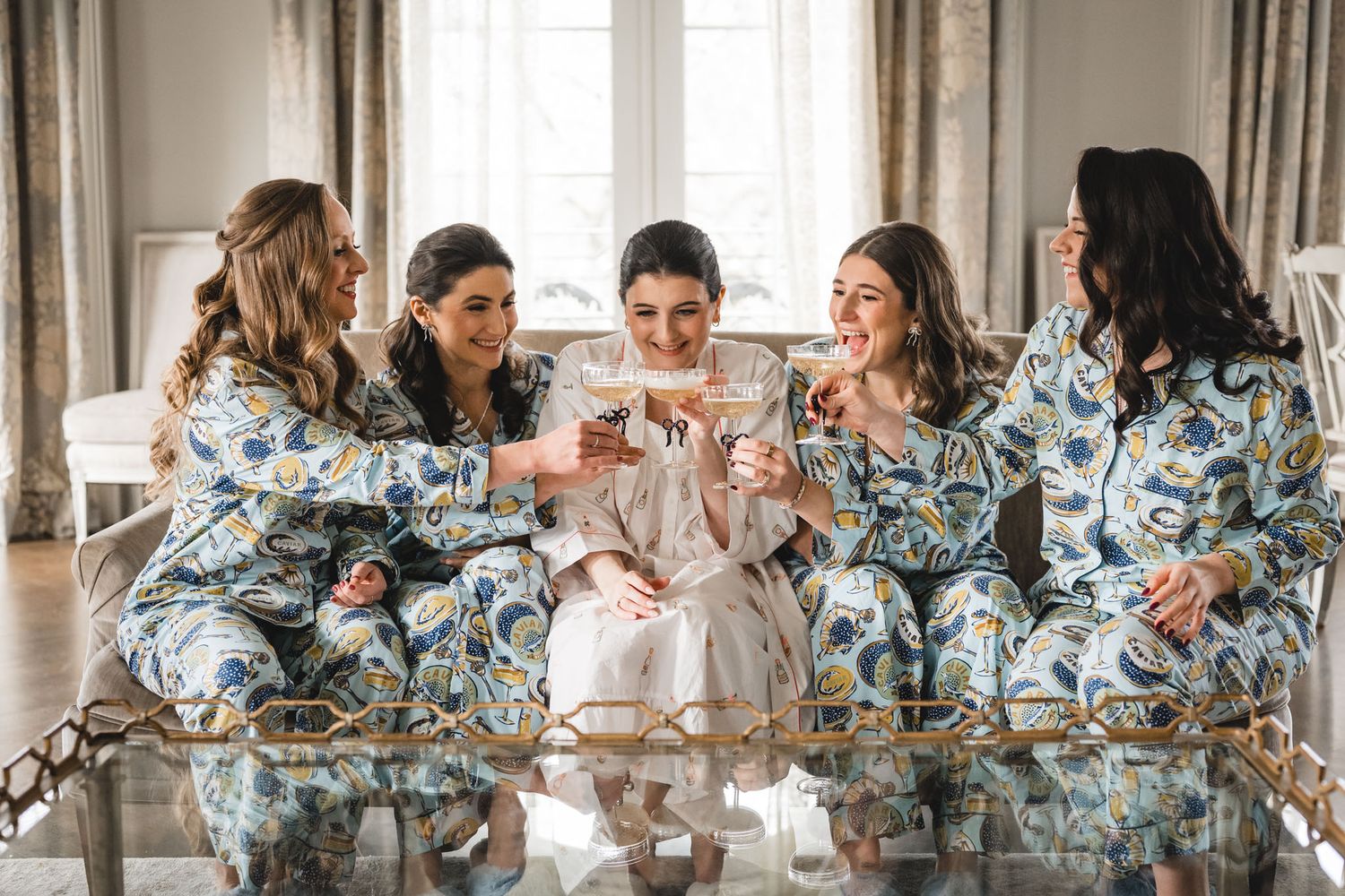 Bridesmaids in matching floral robes toast with champagne glasses at a pre-wedding celebration.