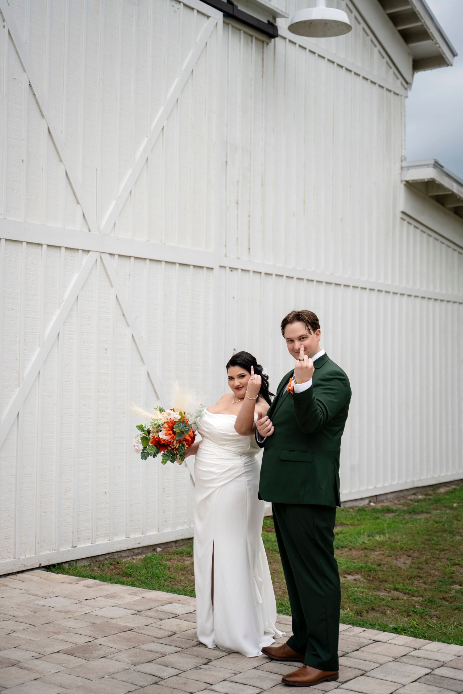 A couple in wedding attire dances playfully in front of a white barn wall.