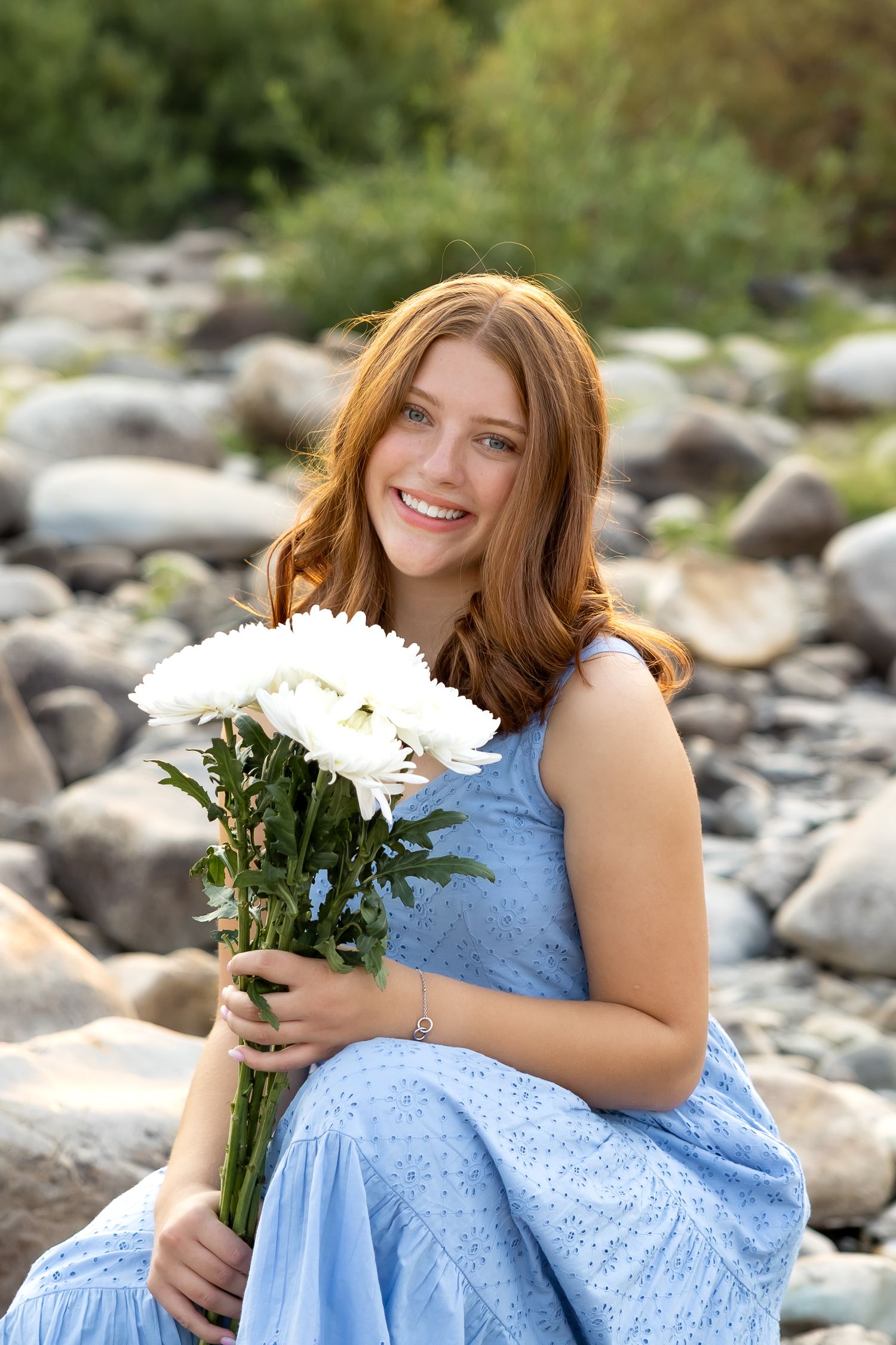 White chrysanthemums complement a blue eyelet dress in a rocky outdoor setting during a photo shoot.