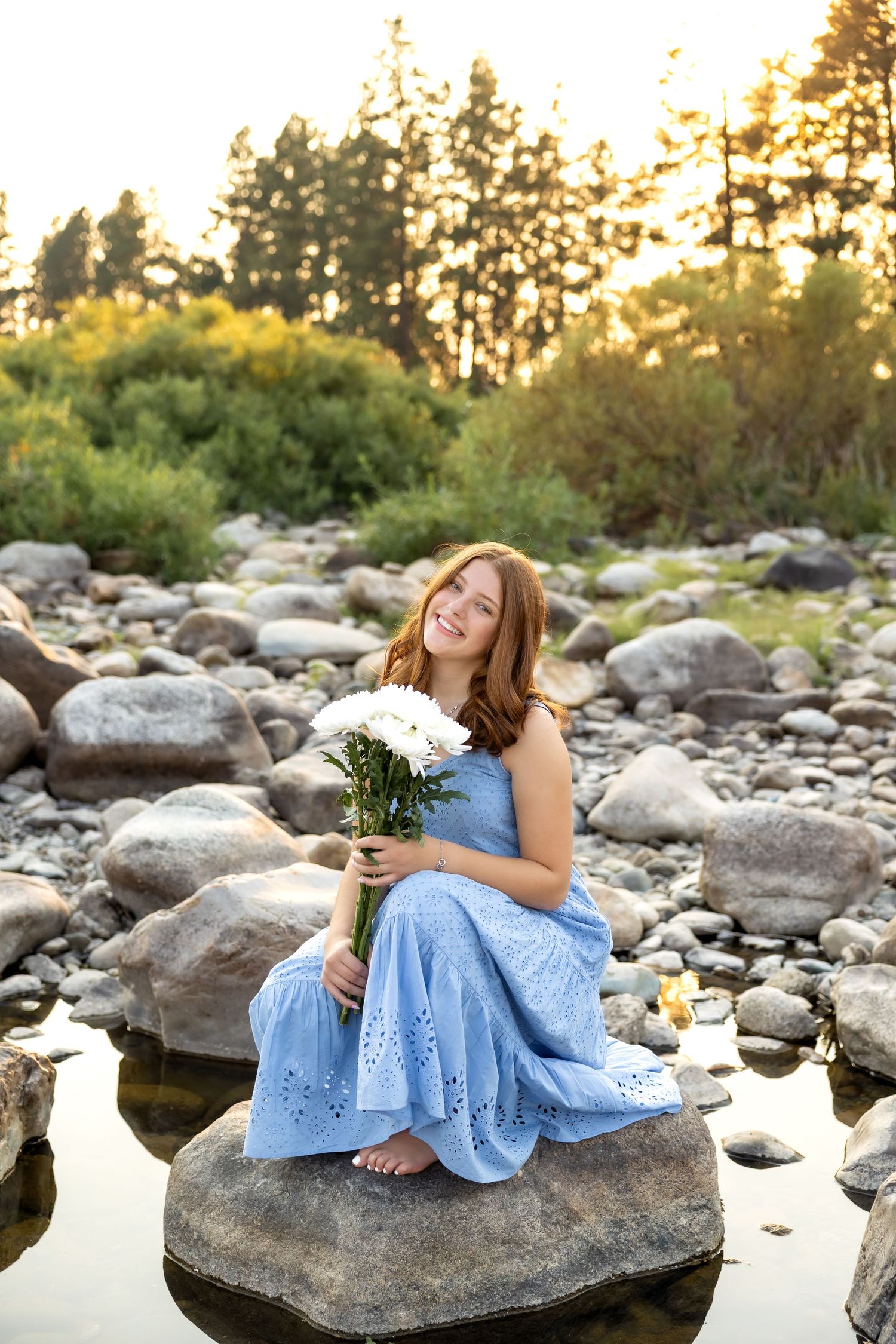 Someone in a blue dress sits on rocks by a stream holding white flowers at sunset.