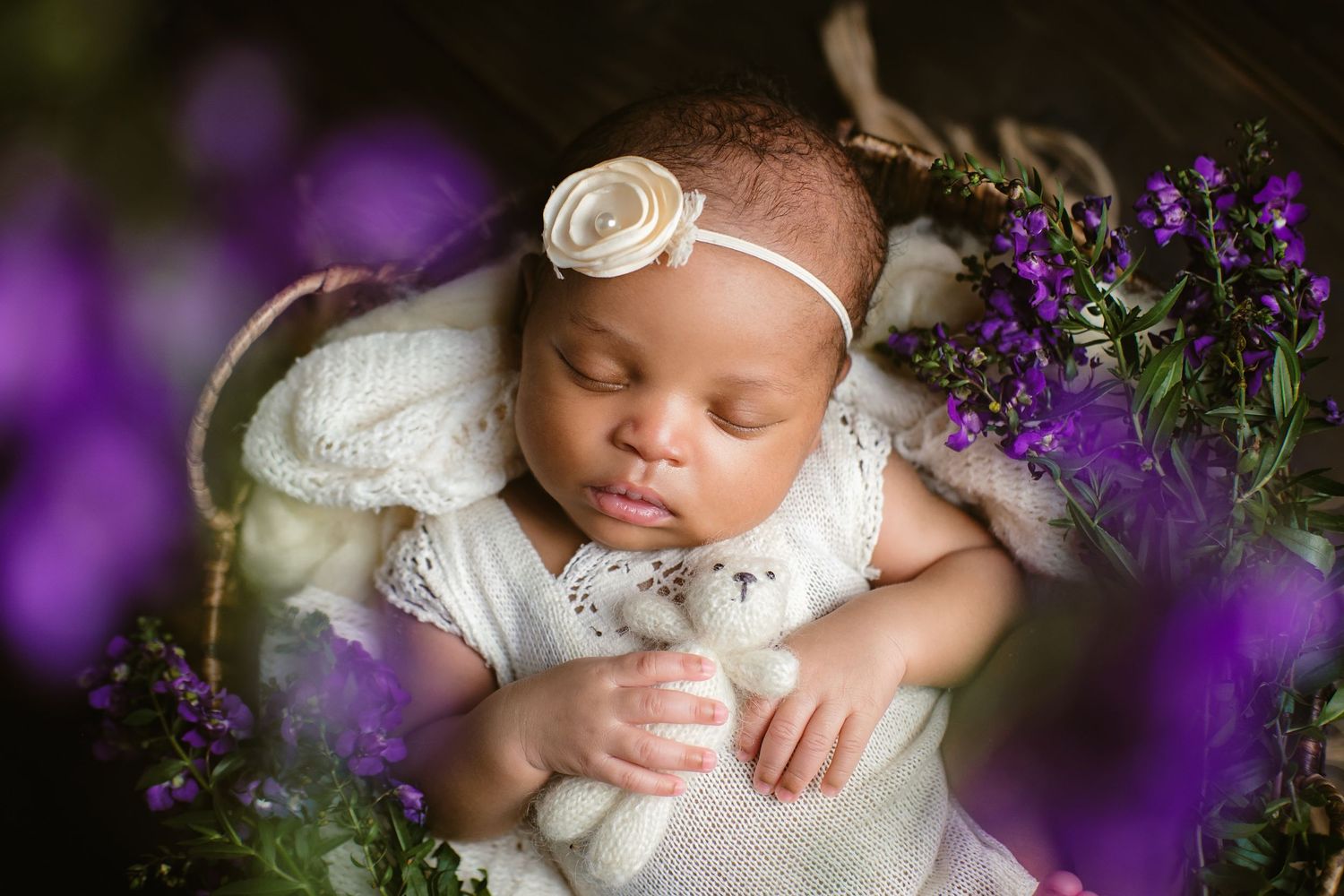 Newborn baby girl holding a teddy bear in a vintage setup surrounded by purple flowers