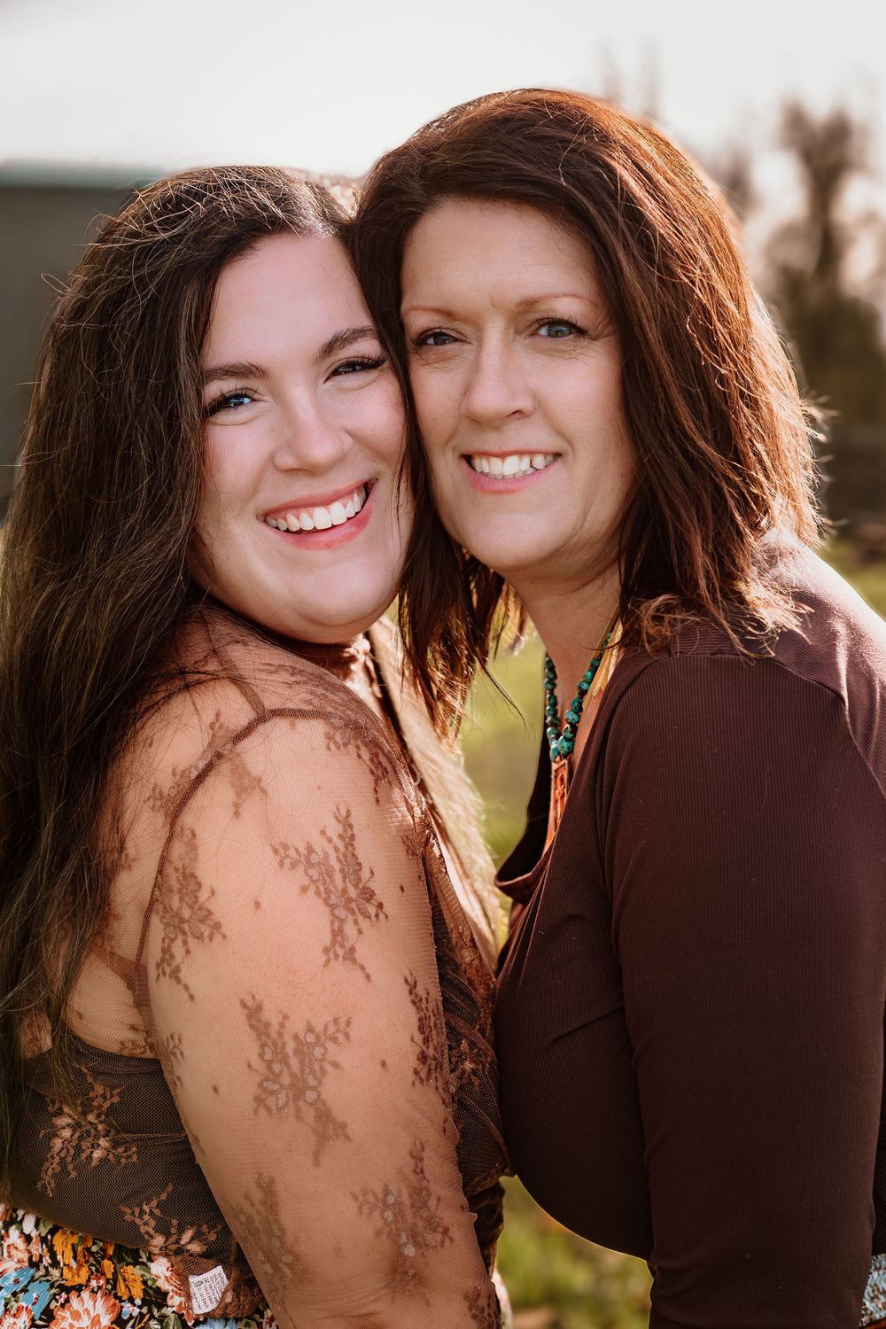 Two smiling women with brown hair pose cheek to cheek outdoors in warm light, wearing floral and brown tops.
