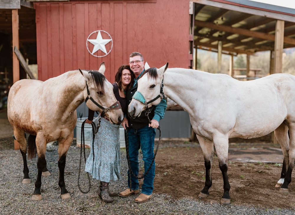 Couple smiling between two horses outside a red barn with a star emblem at a rustic ranch.