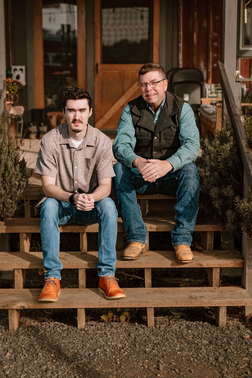 Two men sitting on wooden porch steps of a rustic home, wearing jeans and casual shirts, posing for a portrait.
