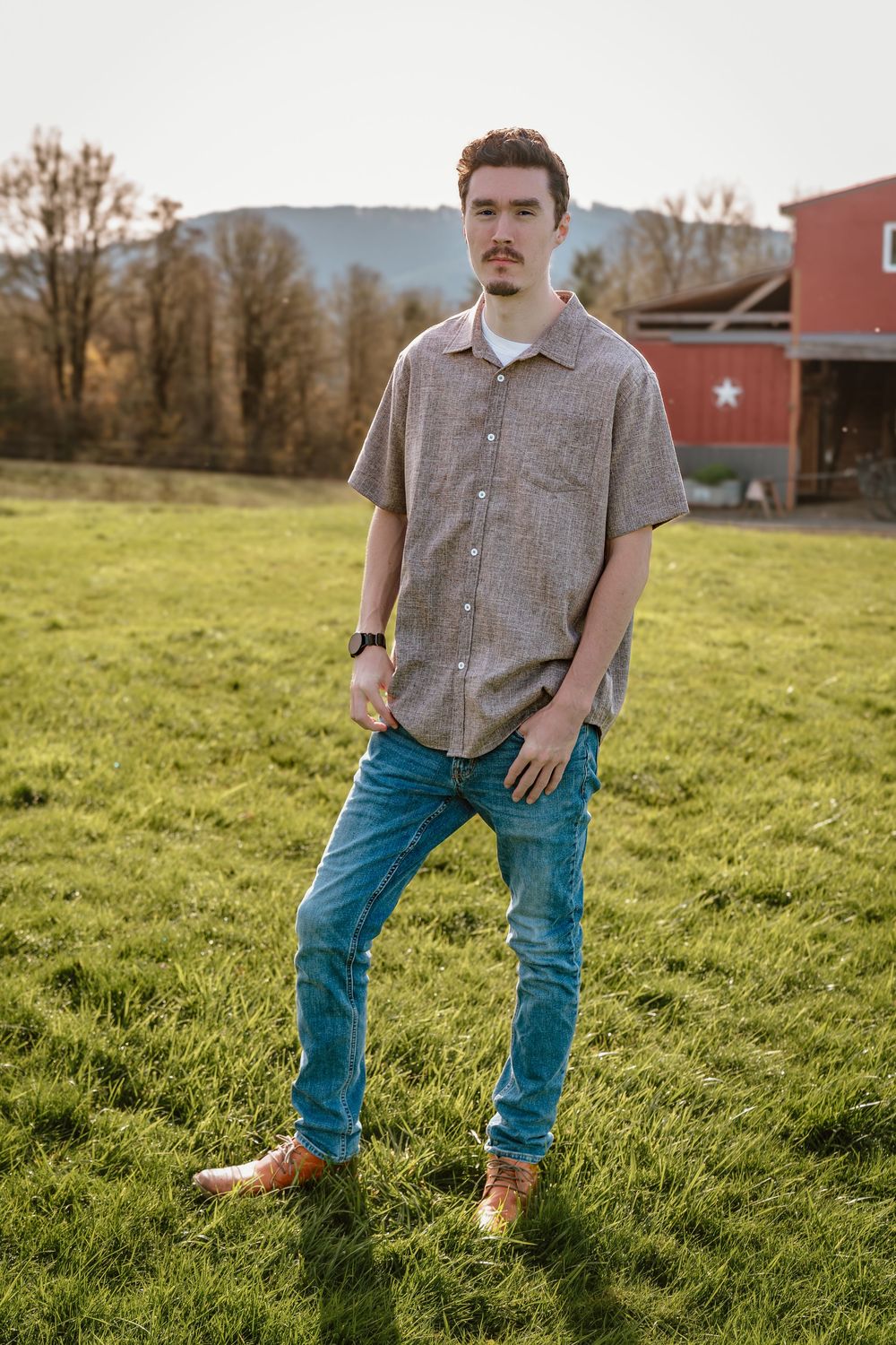 Young man in gray shirt and blue jeans stands on green farm field with red barn and mountains behind.