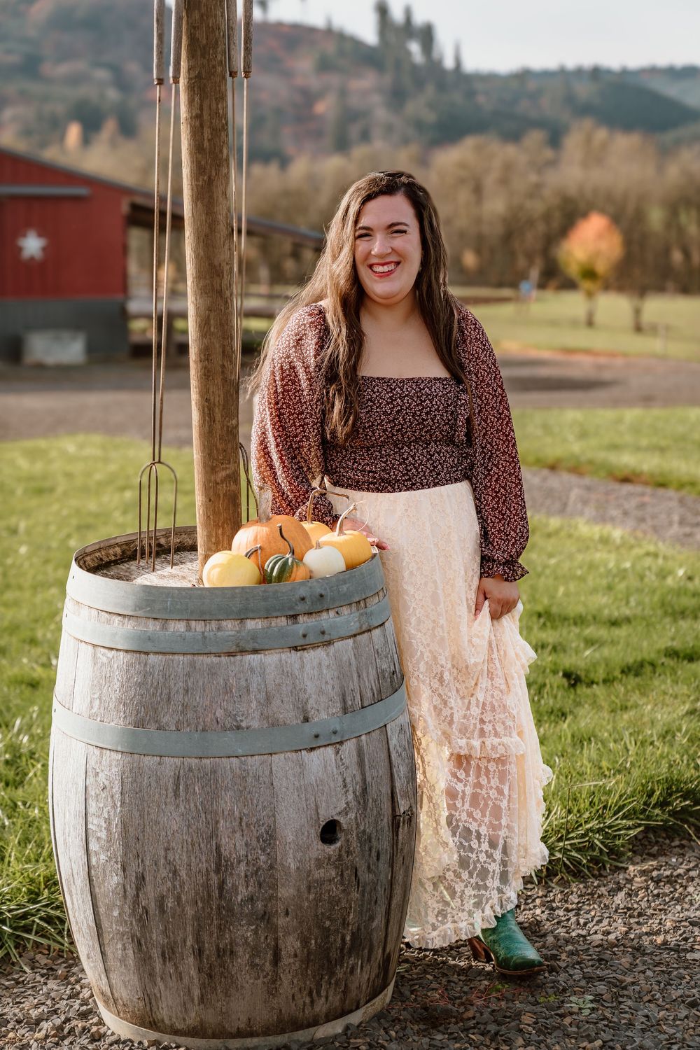 Woman in floral top and lace skirt posing beside a rustic wooden barrel with pumpkins on a fall farm.