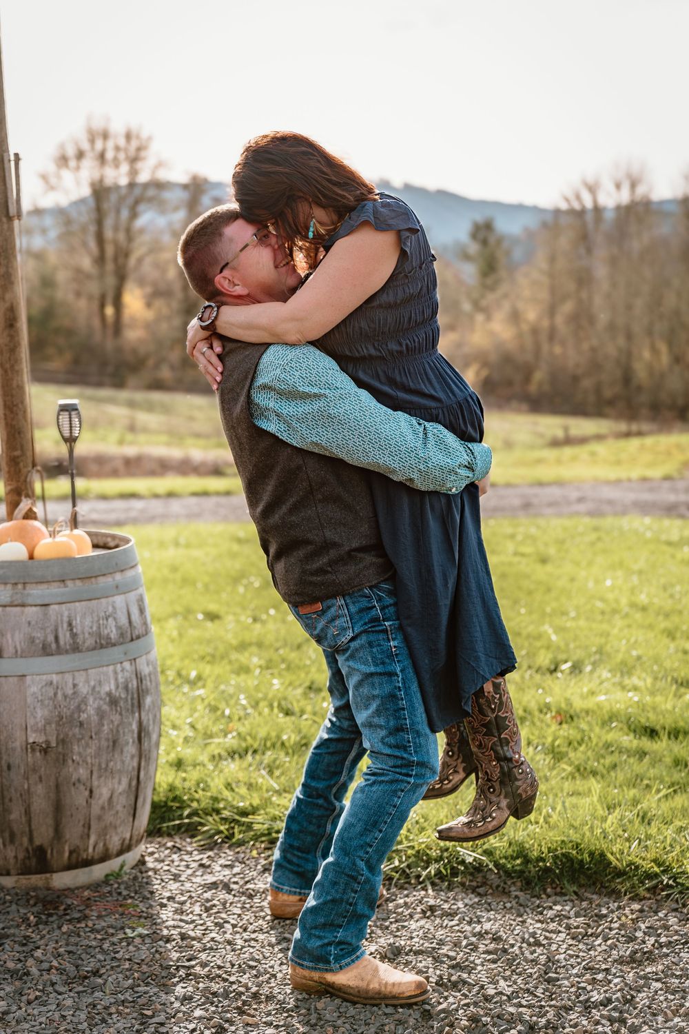 Man lifting woman in romantic embrace outdoors near rustic barrel with pumpkins and green fields in background.
