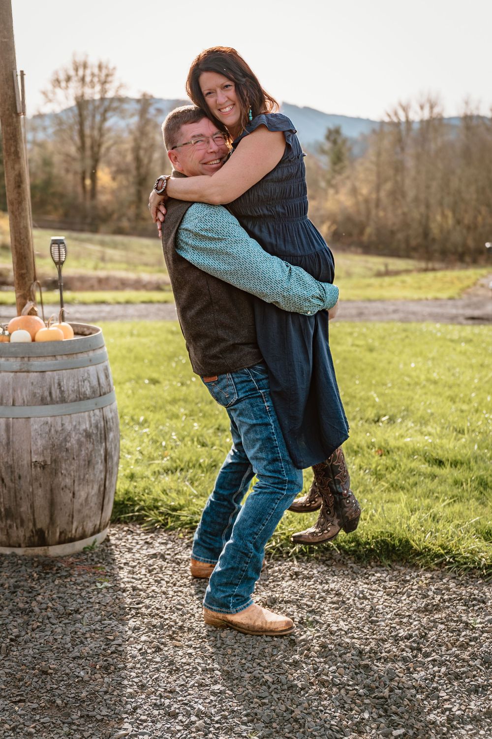 Couple embracing outdoors on a farm, man lifting woman near a wooden barrel with pumpkins, mountains in background.