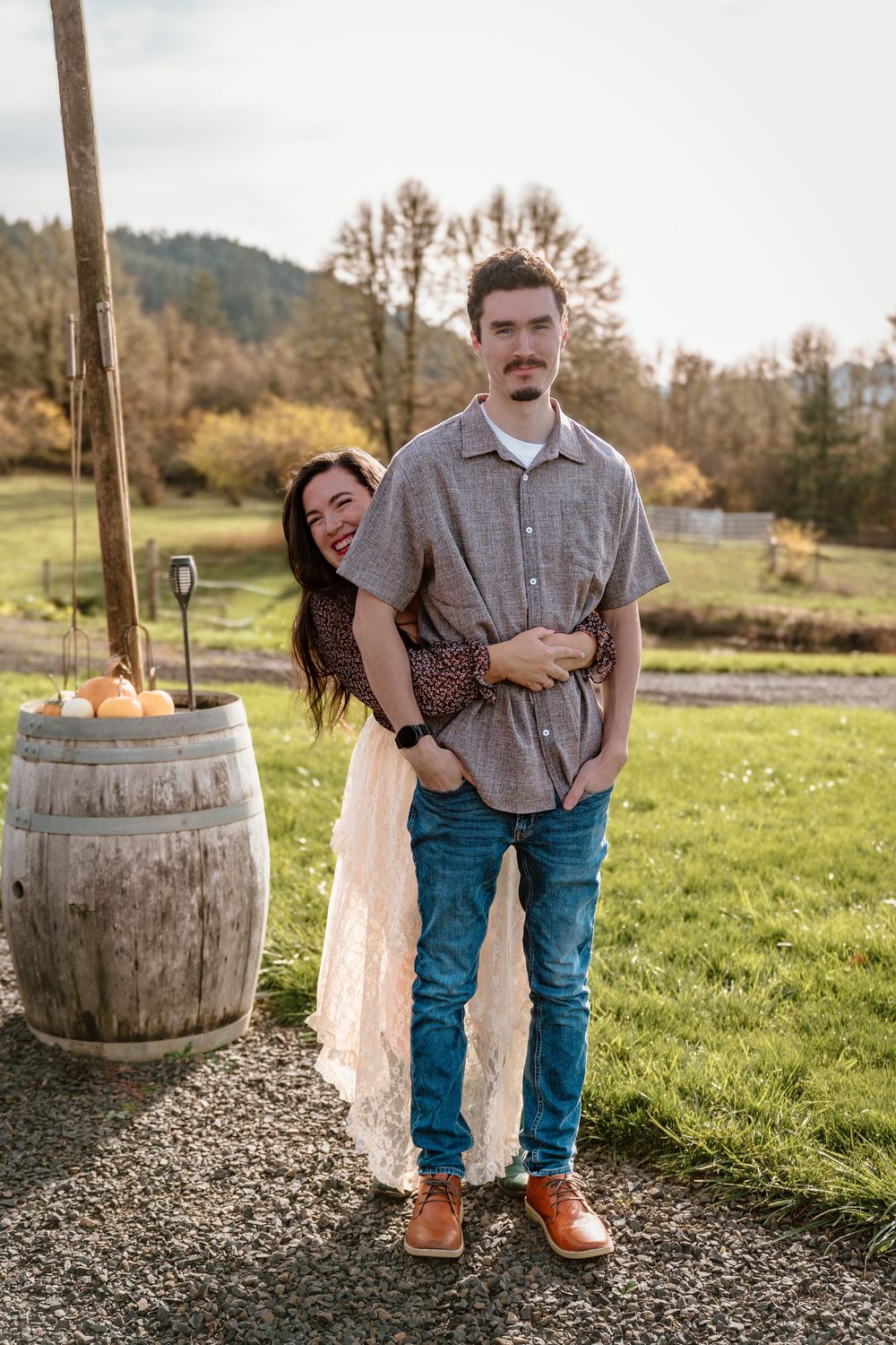 Couple posing outdoors near a rustic wooden barrel with apples, surrounded by green fields and trees in autumn.