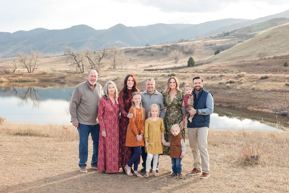 Family group poses together in casual attire against scenic mountain backdrop and lake on autumn day.