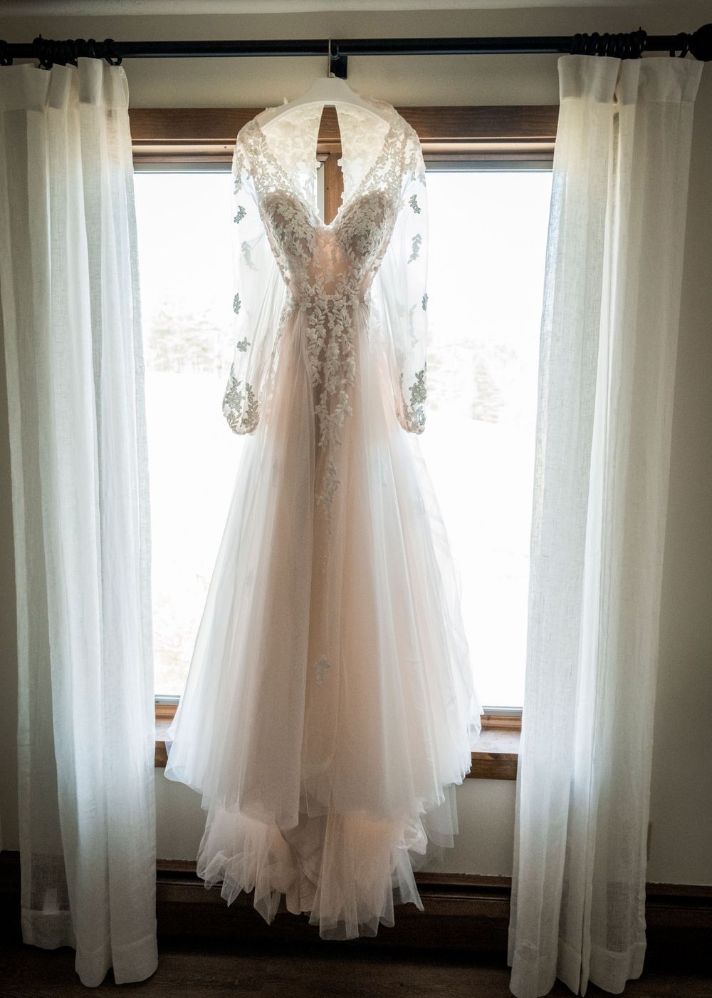 Elegant beaded lace wedding gown with tulle skirt and sheer sleeves hanging in a window.