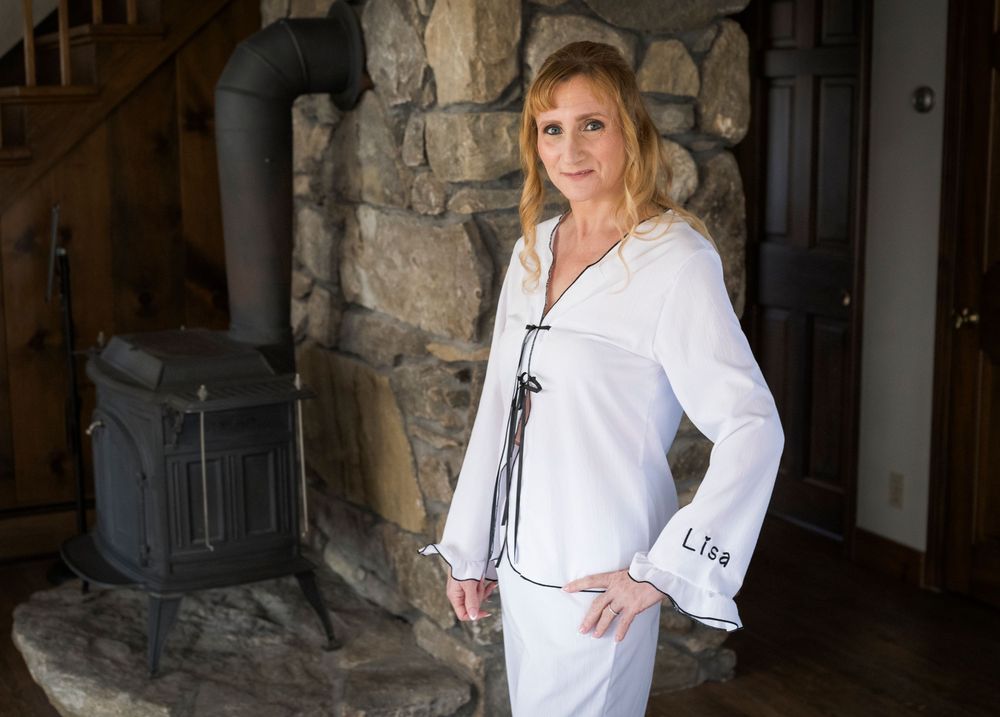 Woman in monogrammed white pajamas posing by a stone fireplace with wood stove.