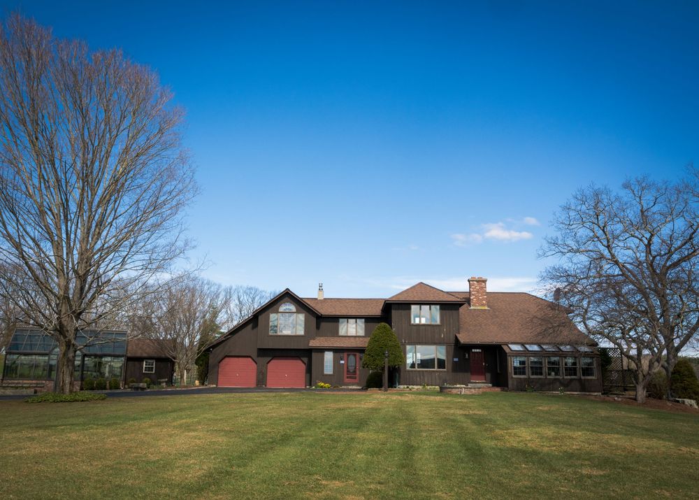 Large brown suburban home with red garage doors, expansive green lawn, and bare trees under a bright blue sky.