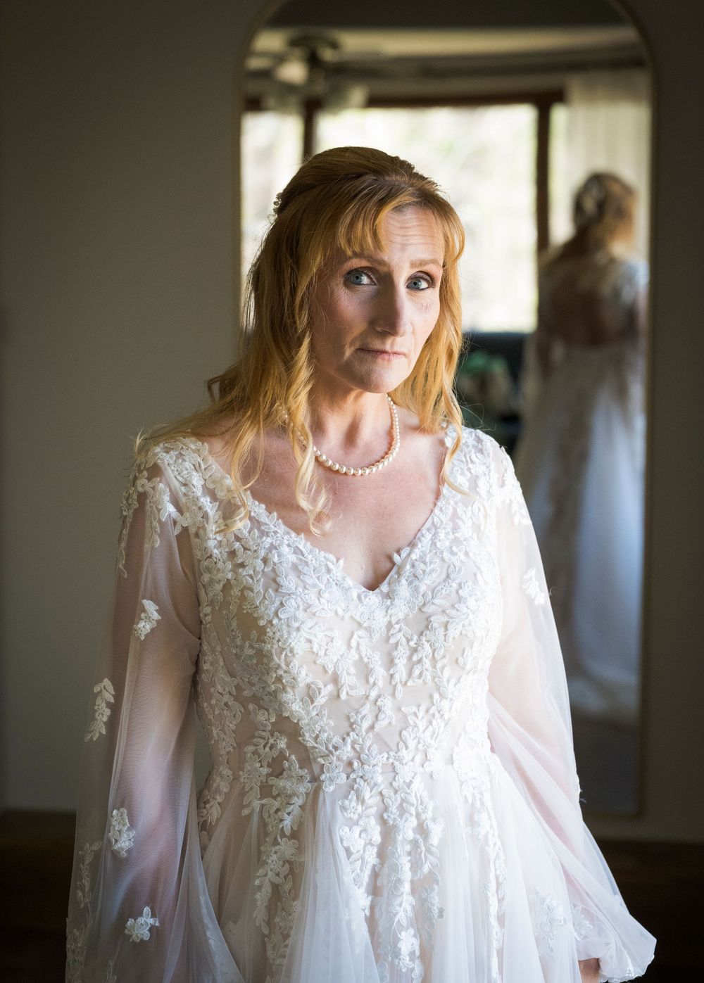 Bride in lace wedding gown with sheer sleeves and pearl necklace stands indoors with her reflection visible in background.