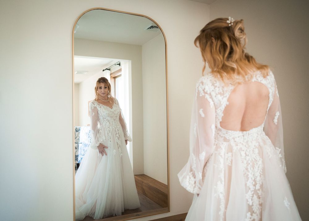 Bride in elegant lace wedding gown with open back admiring her reflection in a tall arched mirror.