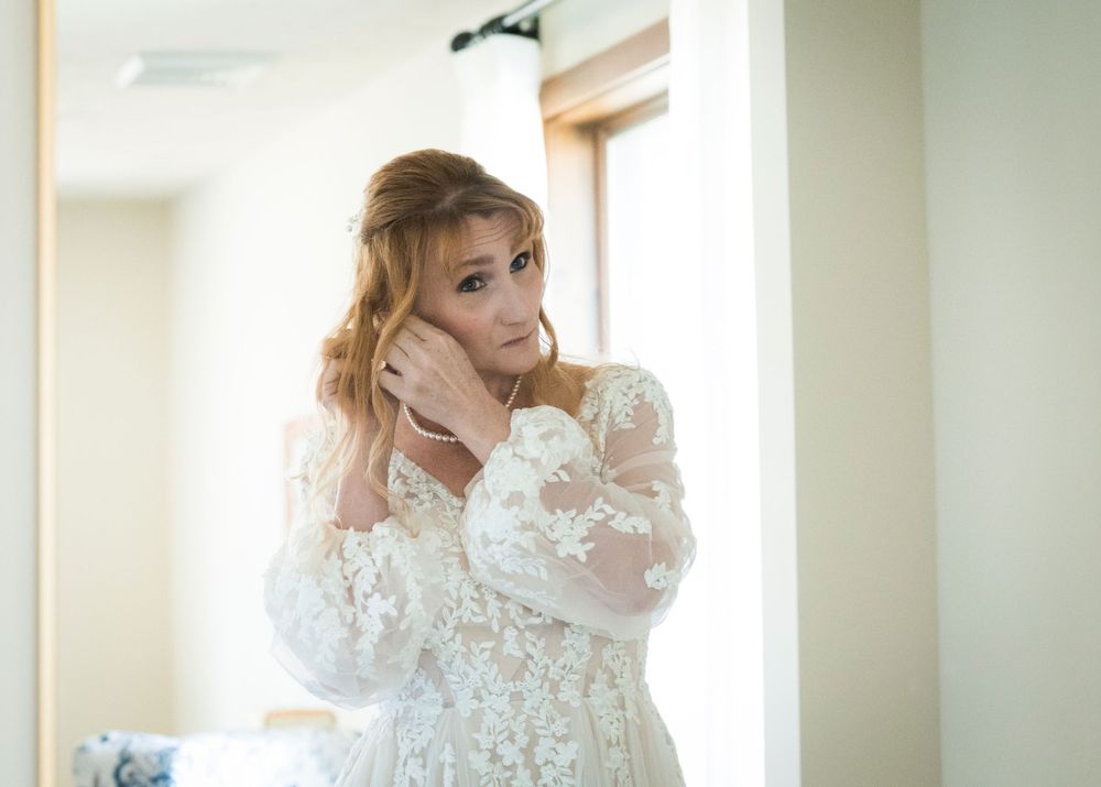 Bride in lace wedding gown adjusting earrings while getting ready in front of mirror.