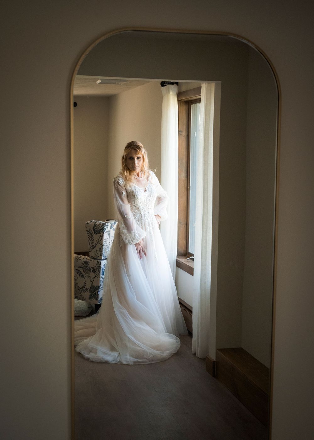 Bride in lace long-sleeve wedding gown standing by window, reflected in arched gold mirror.