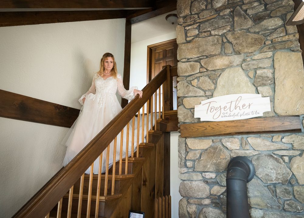 Bride in lace wedding gown descending rustic wooden staircase beside stone fireplace wall with Together sign.