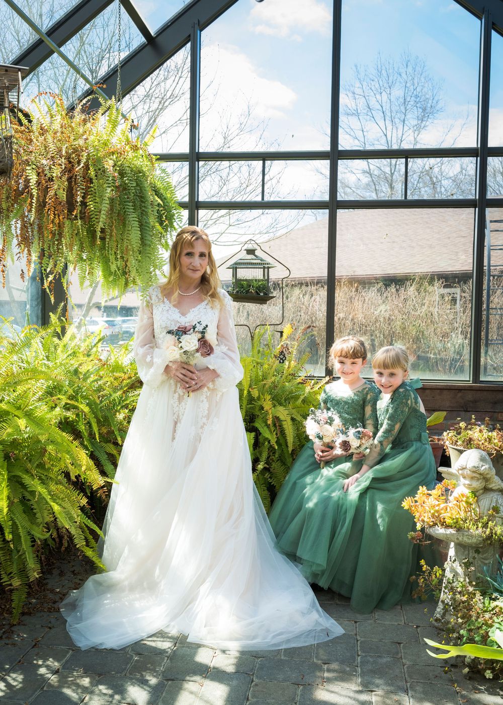 Bride in white lace gown poses with two flower girls in green dresses inside a sunlit greenhouse.