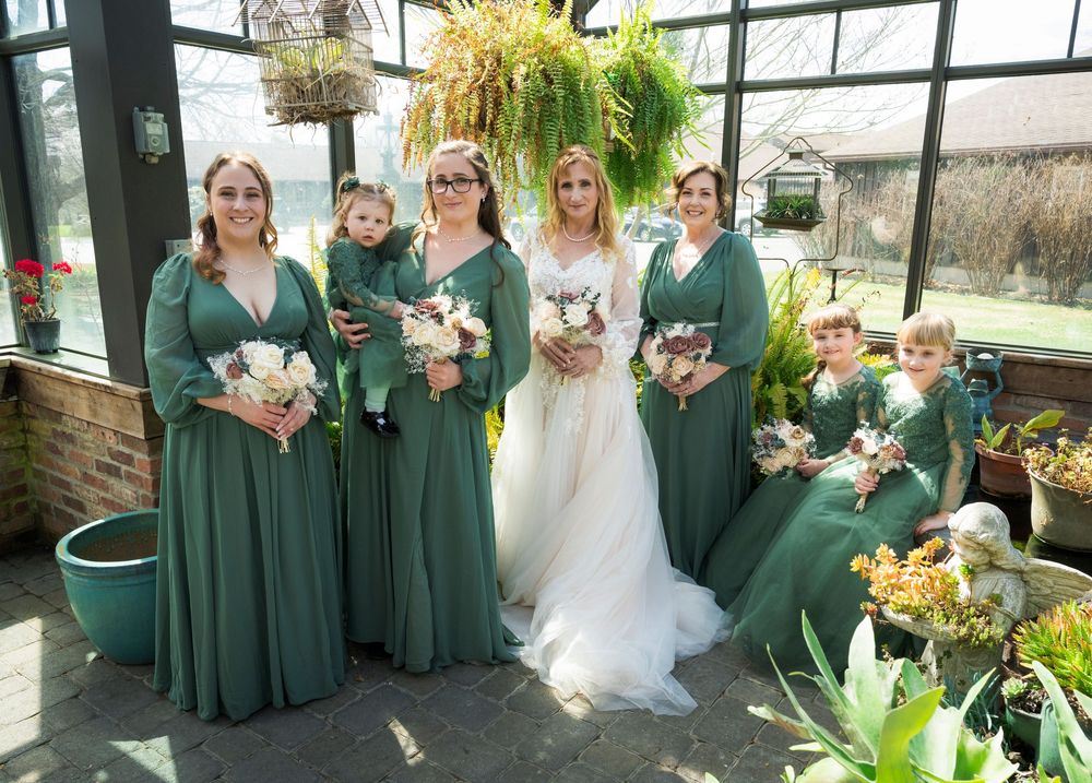 Bride in white gown poses with bridesmaids in emerald green dresses and flower girls in a greenhouse wedding venue.