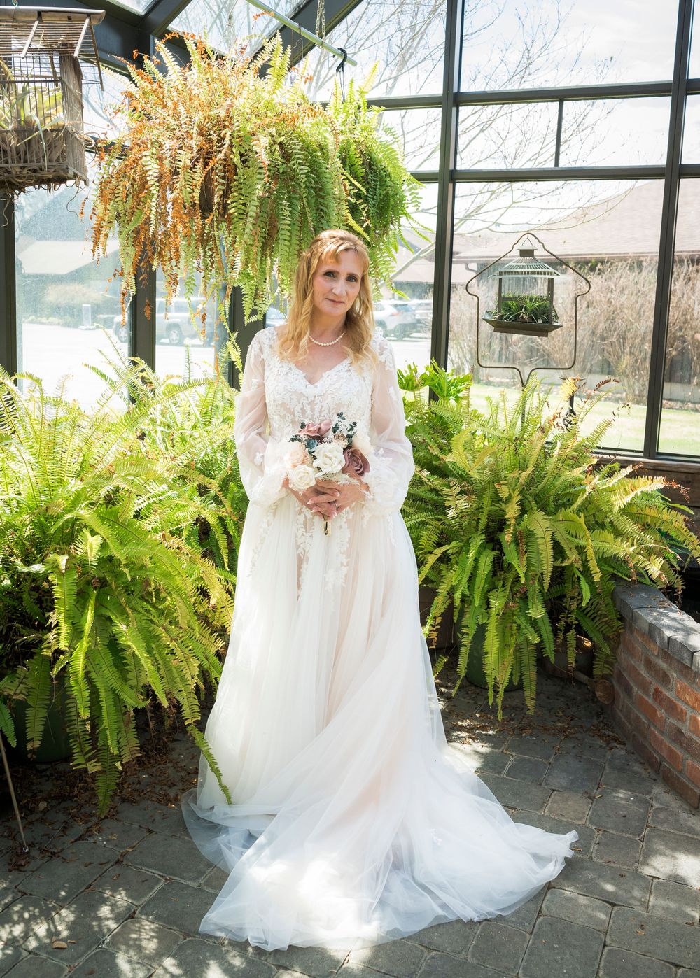 Bride in lace wedding gown with long sleeves holding bouquet in a sunlit greenhouse with lush ferns.